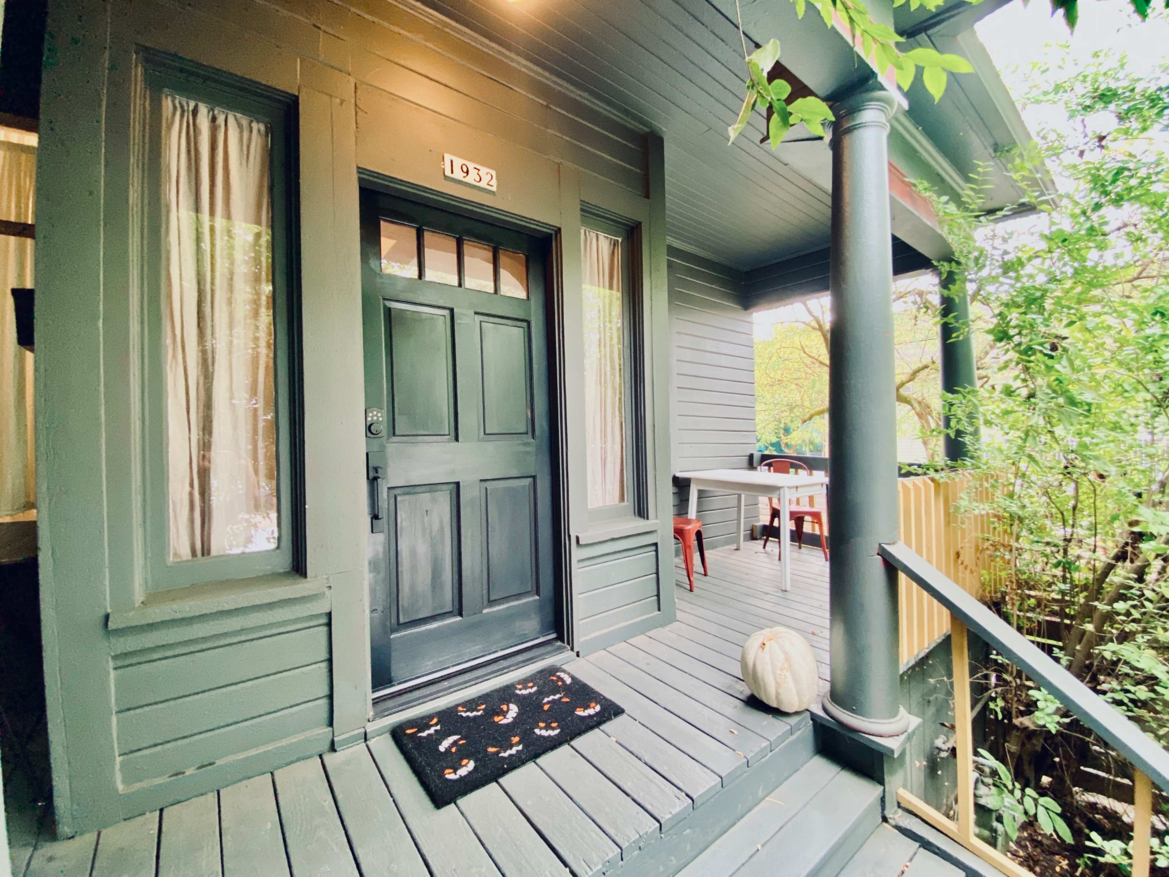 The image shows the entrance of a house with a dark green door, a welcome mat, and a small table and chairs nearby, surrounded by greenery.
