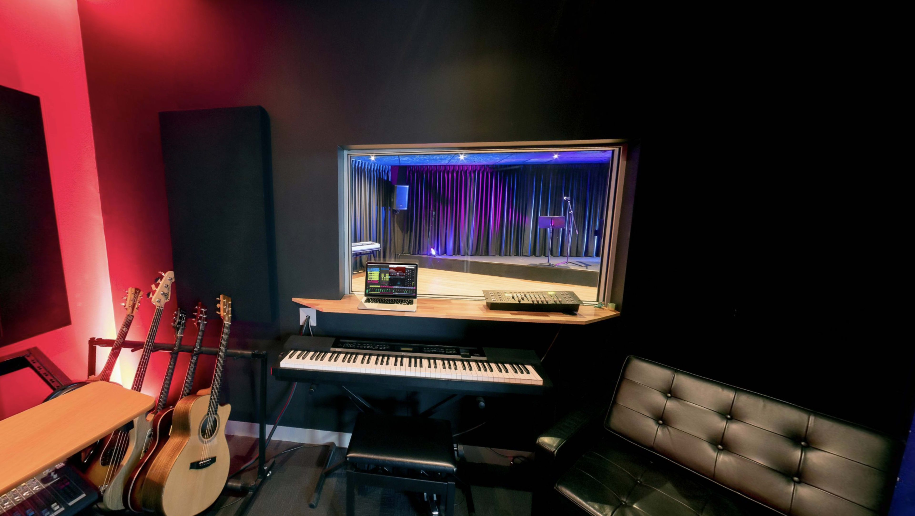 The image shows a recording studio featuring a piano, music production equipment, and guitars, with a window overlooking a stage area lit by purple and blue lights.