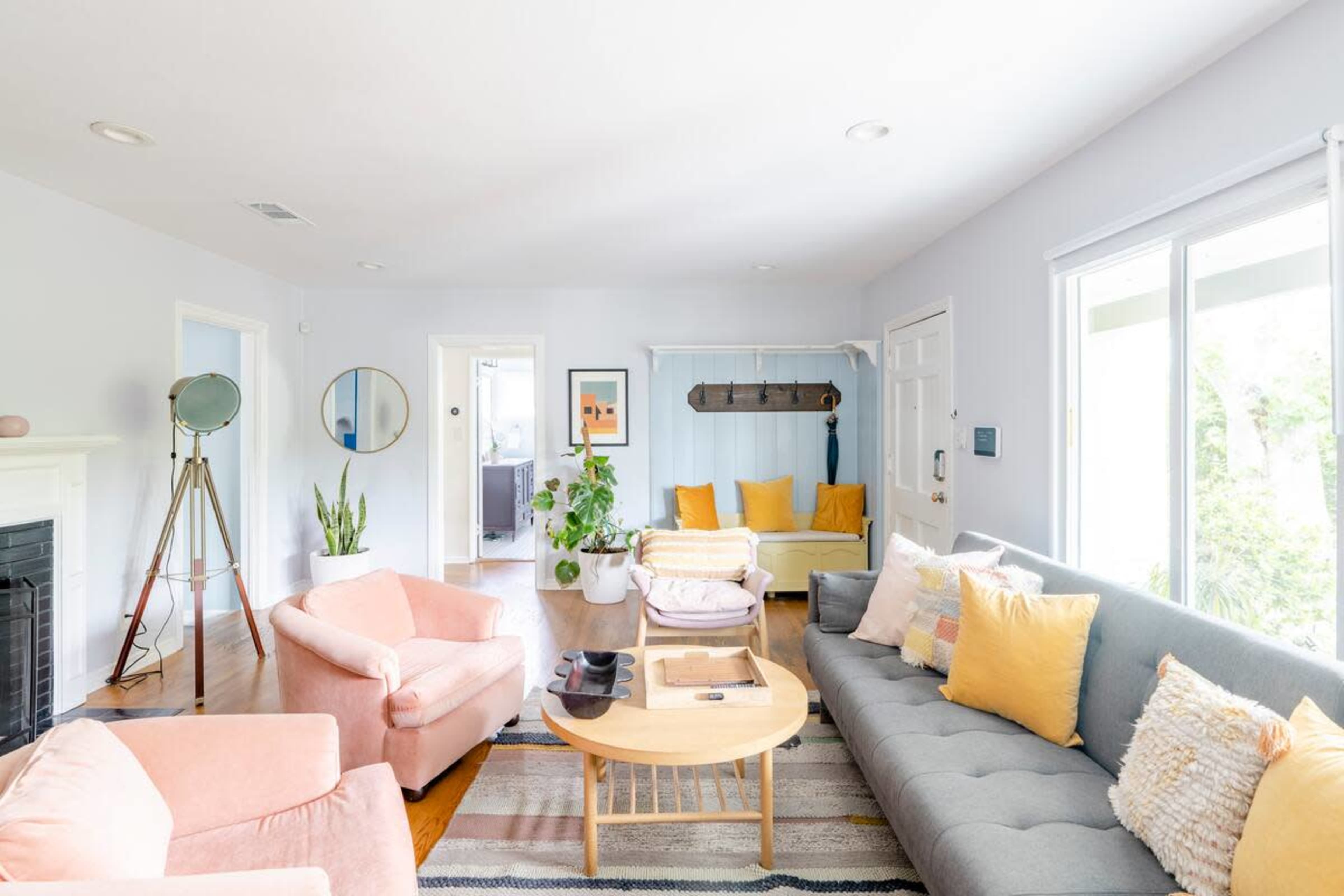 The living room features a light color palette with a gray sofa, pink armchairs, and a wooden coffee table, along with a yellow accent seat and a plant near a large window.