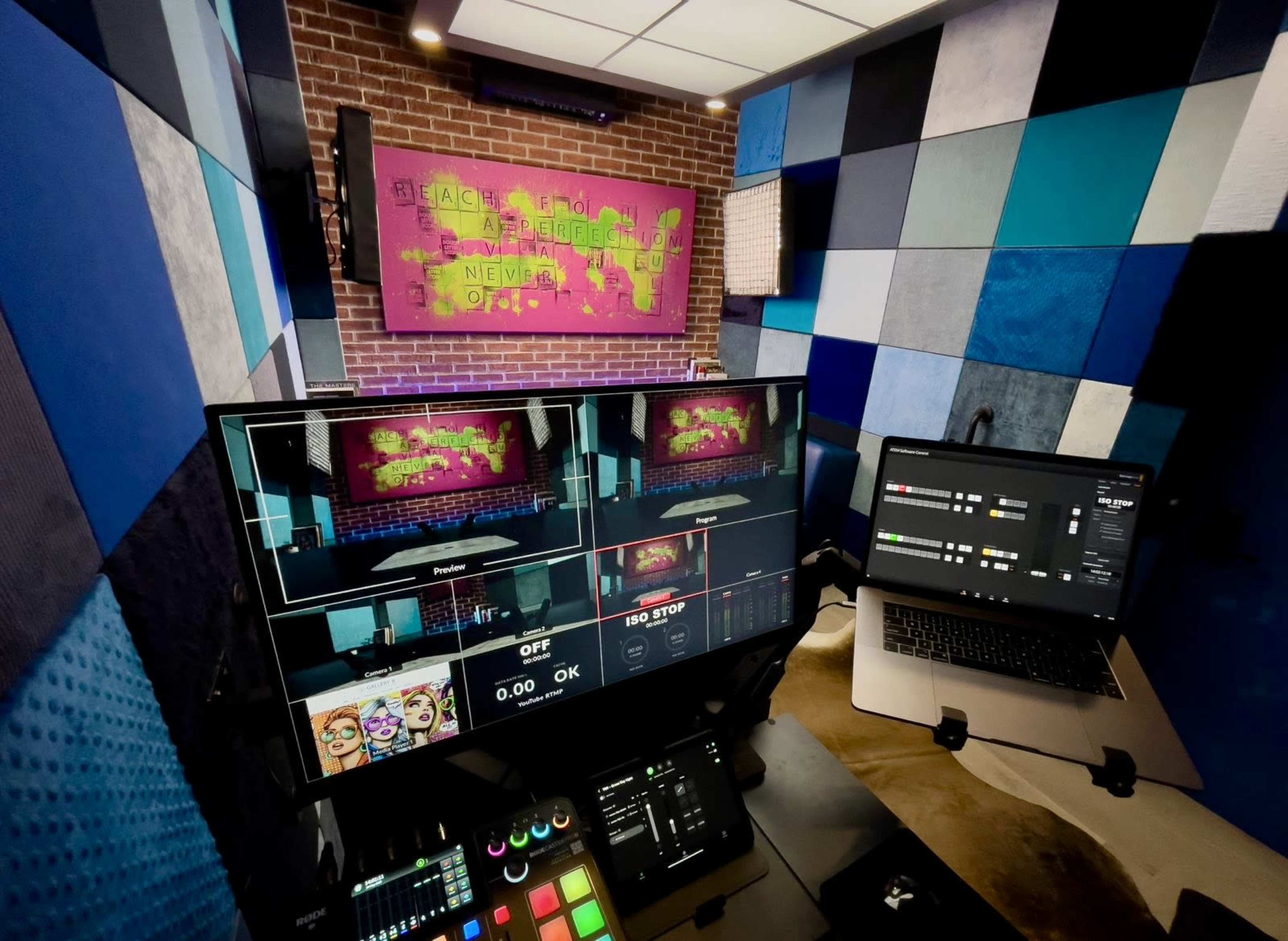 A control room setup featuring multiple screens, a laptop, and a colorful blue and gray soundproof backdrop with a brick wall displaying artwork.