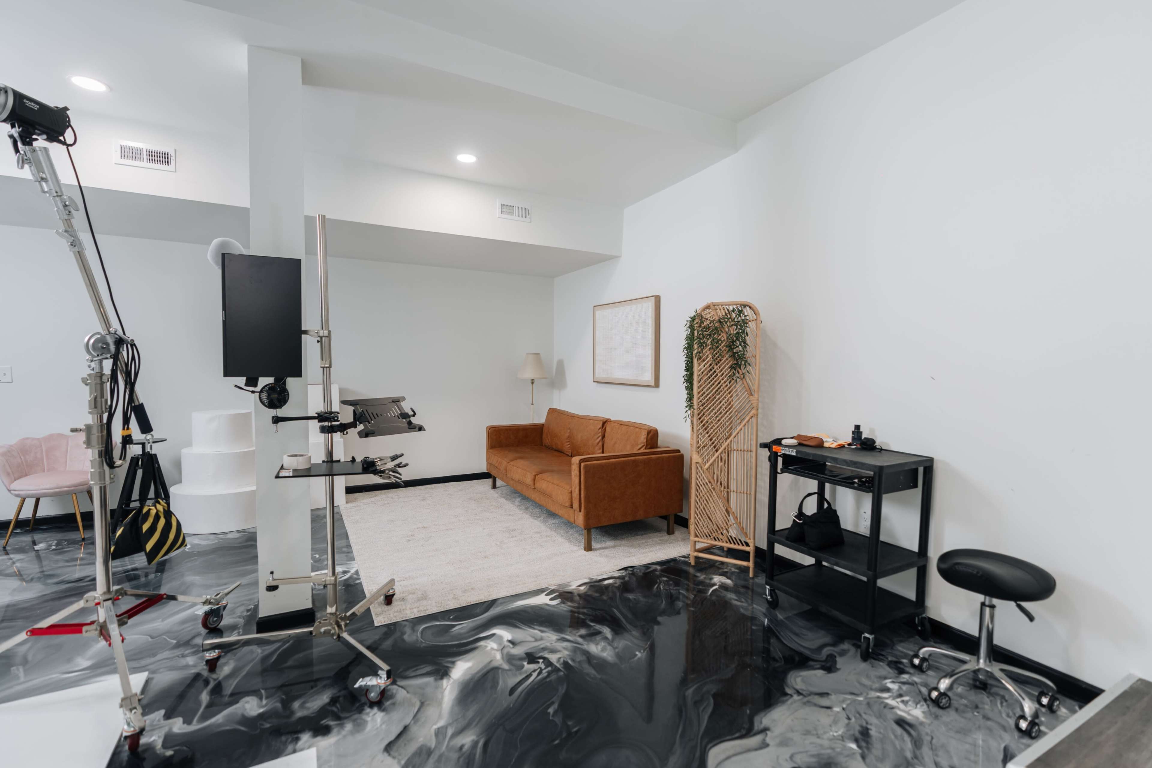 The image shows a modern studio space with a brown sofa, a small black cart, and equipment on stands against a white wall.