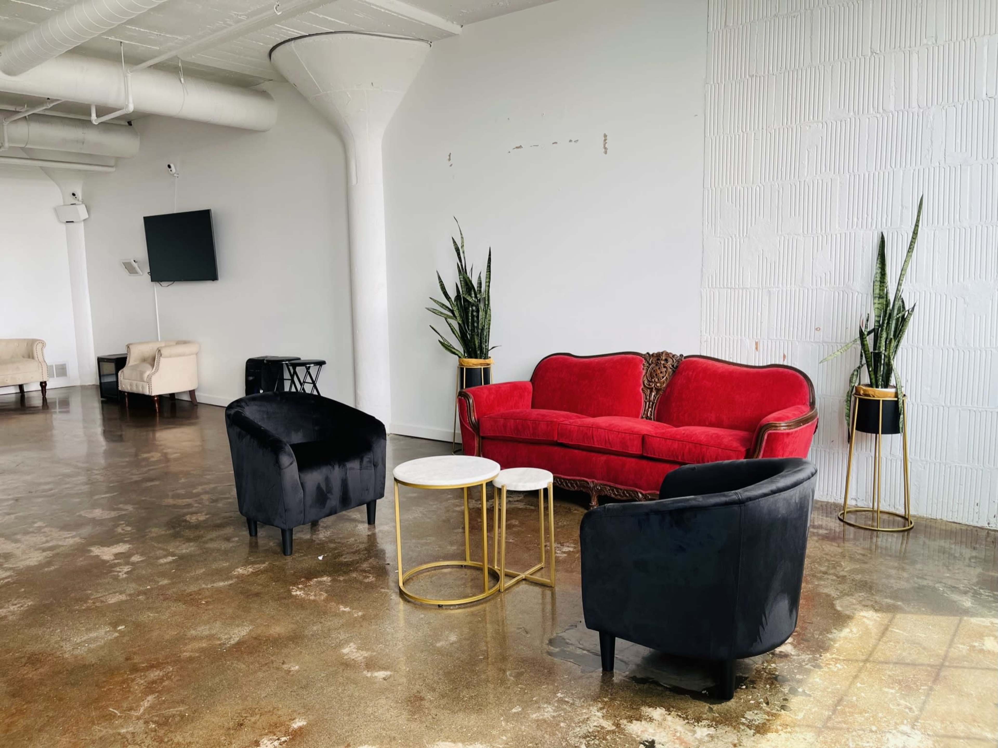 A minimalist interior space featuring a red couch, two black chairs, a white coffee table with a gold base, and potted plants against a white wall.