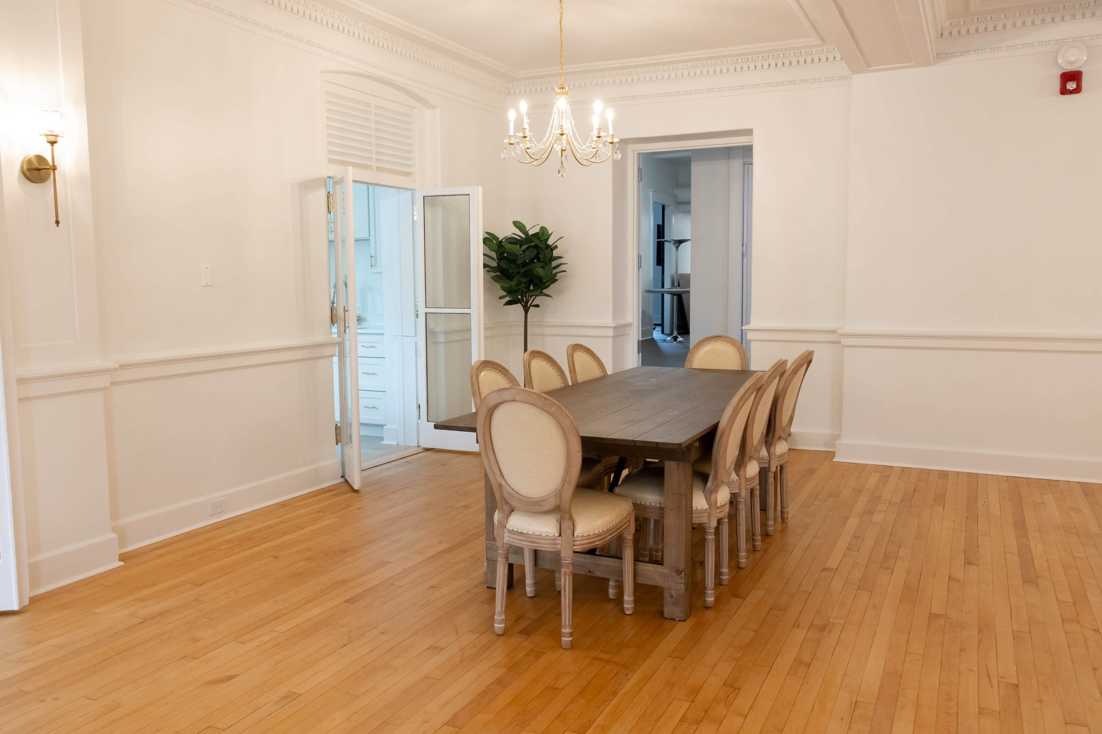 A spacious dining room features a wooden table surrounded by eight chairs, with a chandelier overhead and a potted plant near an open door.