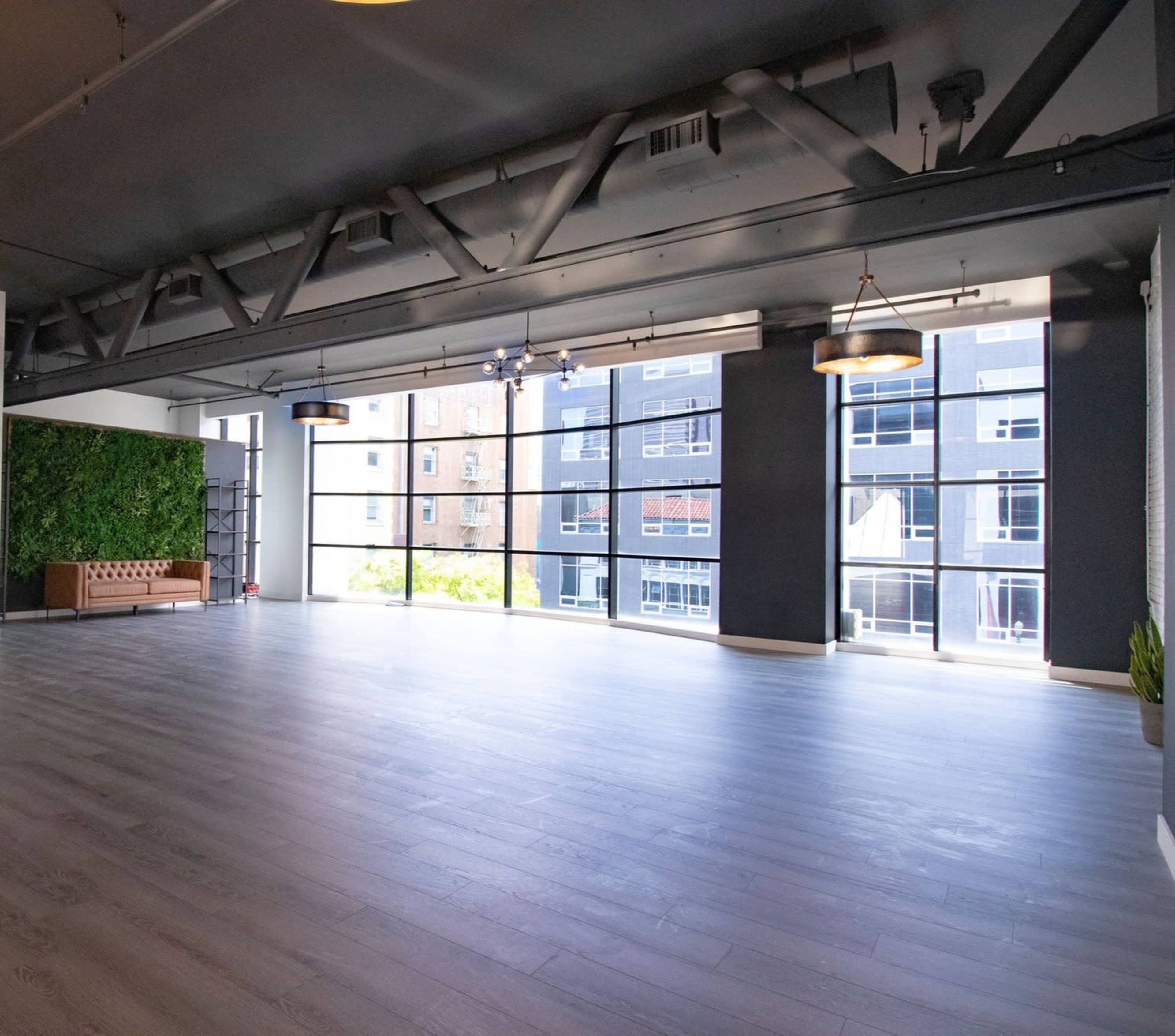 A spacious room with large windows, gray walls, and wood flooring is illuminated by overhead lights.