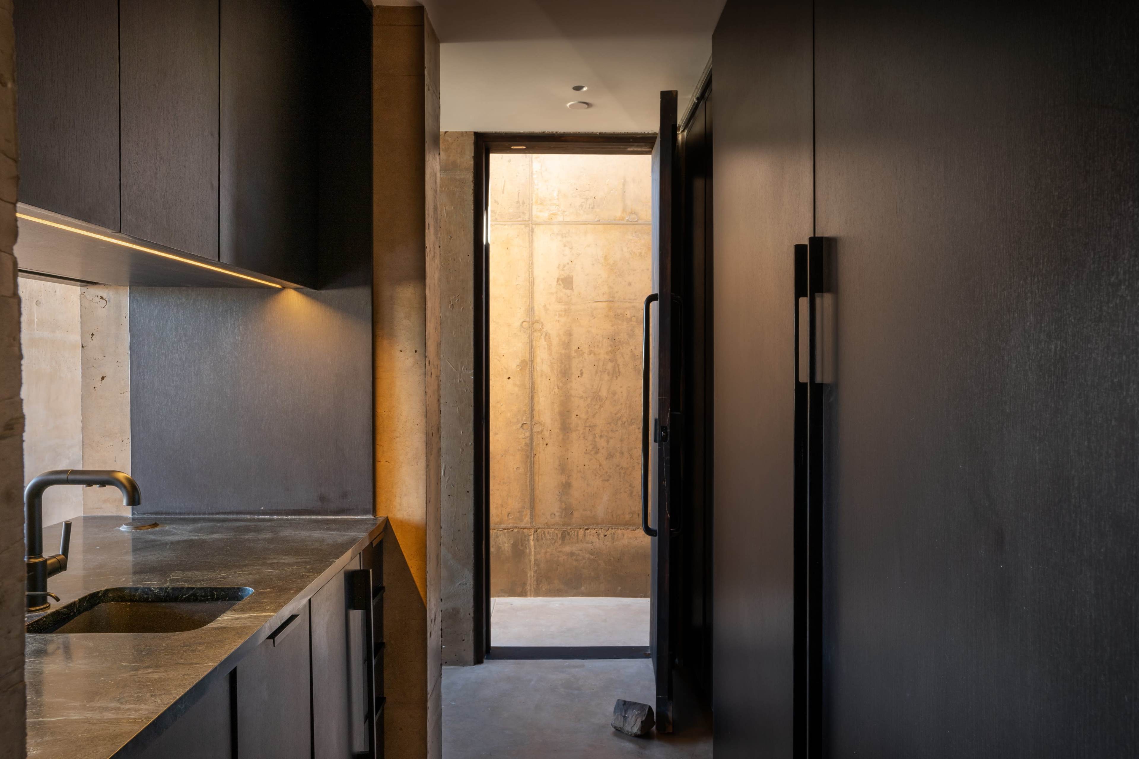 A narrow kitchen space with dark cabinetry and a doorway leading to a light-filled hallway with concrete walls.