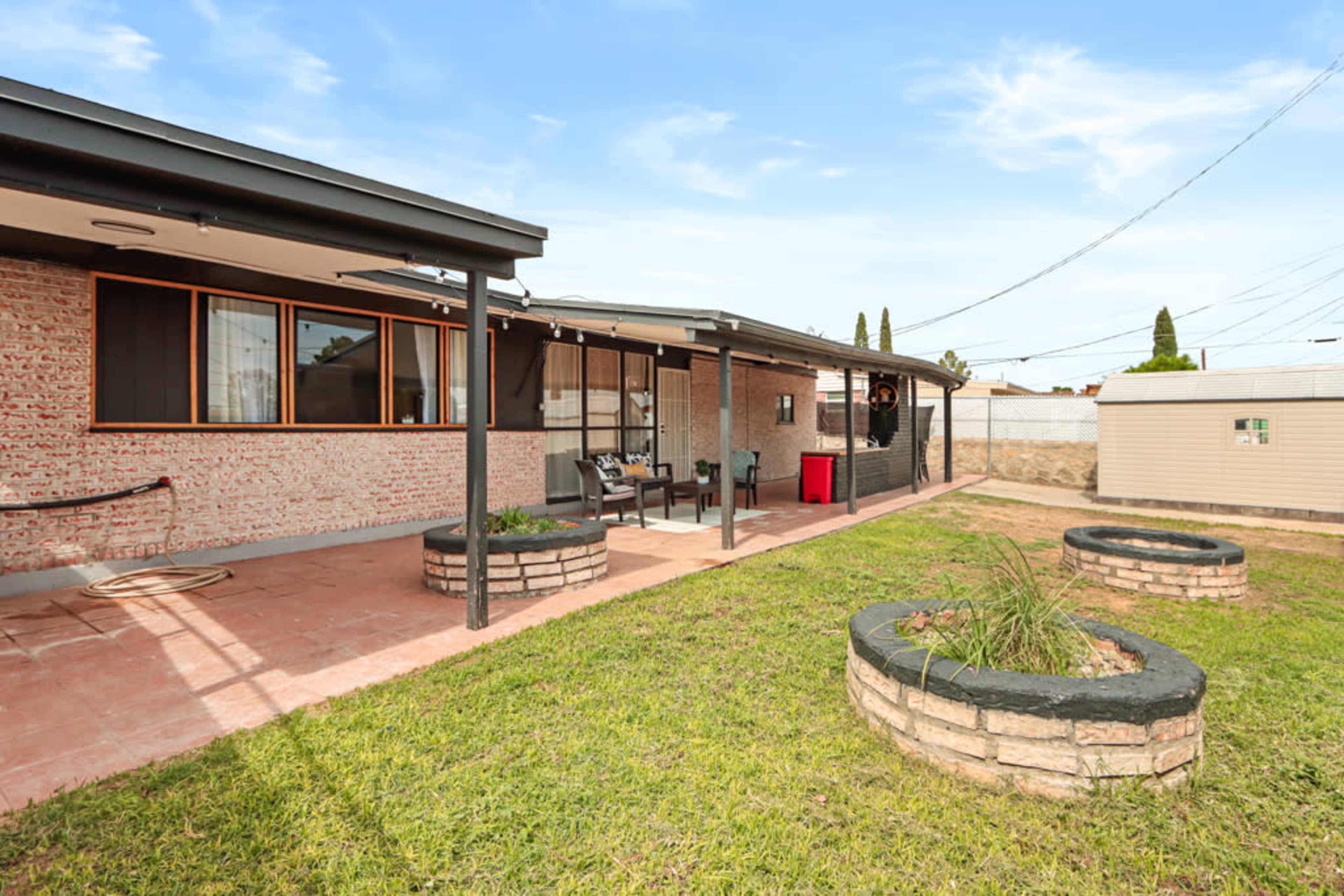 A single-story house with a stone patio and a well-maintained lawn, featuring circular flower beds and a shaded porch area.