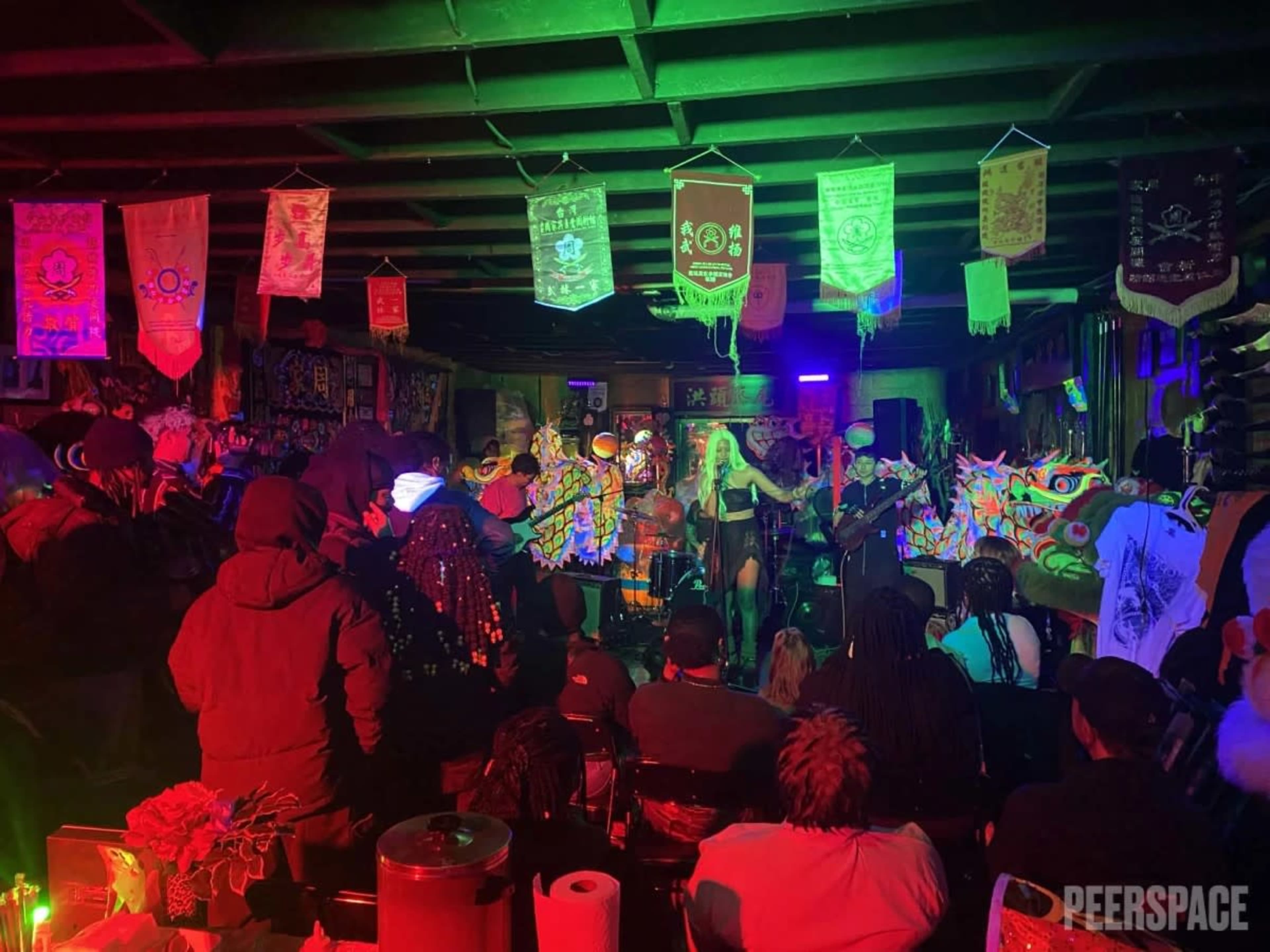 A live music performance takes place in a dimly lit venue filled with colorful decorations and an audience seated in front of the stage.