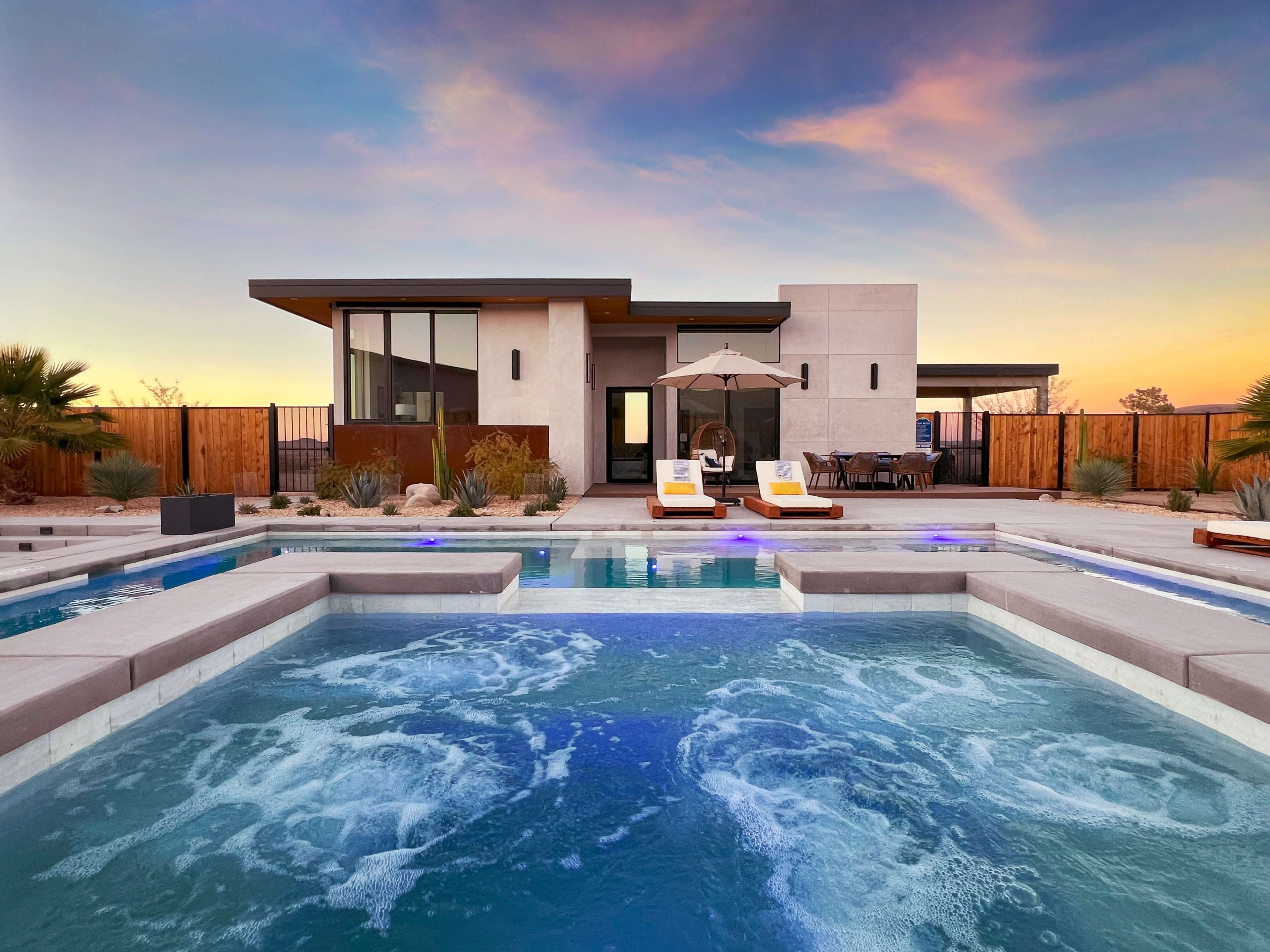A modern house with large windows overlooks a swimming pool with a hot tub, surrounded by desert landscaping and seating areas at sunset.
