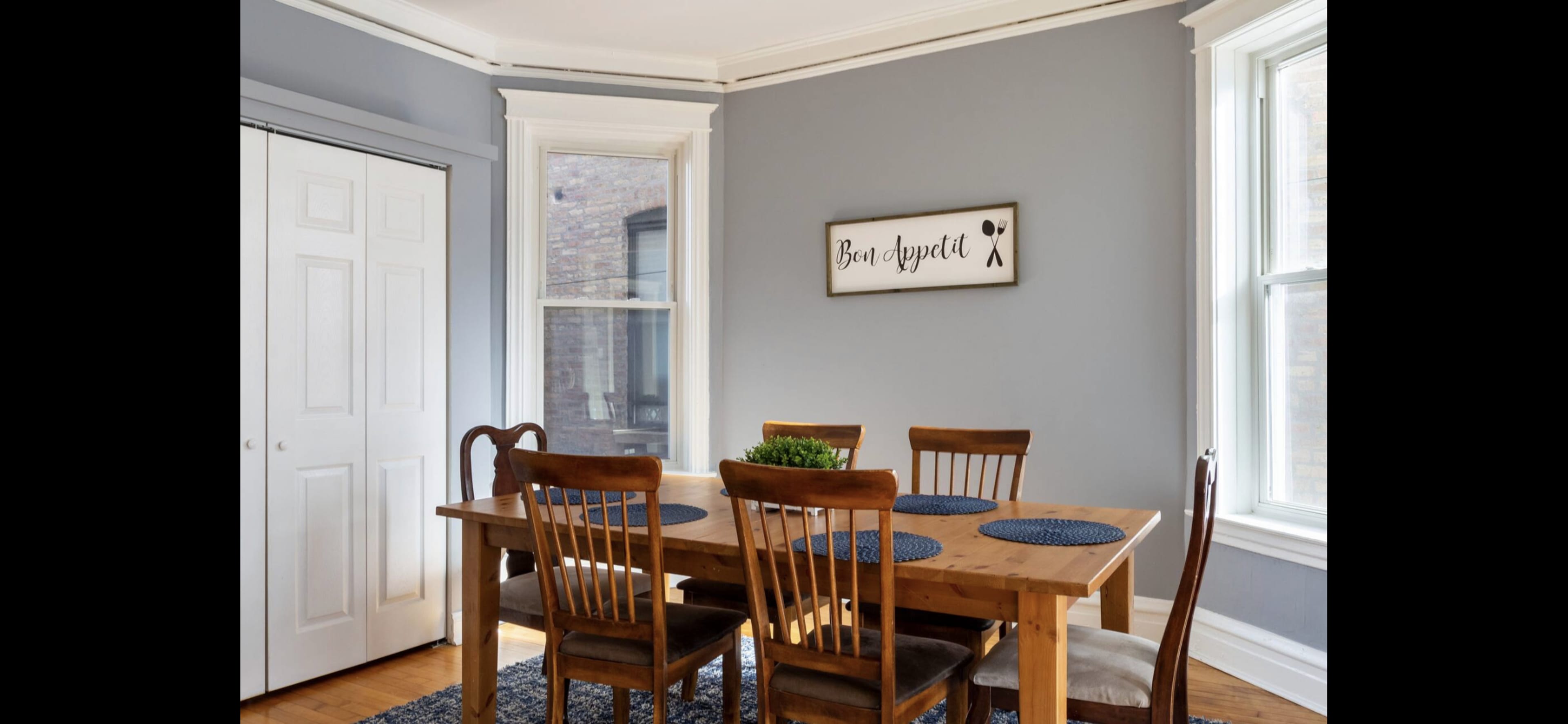 A dining area features a wooden table with chairs, blue placemats, and a wall-mounted sign, set against gray walls and large windows.