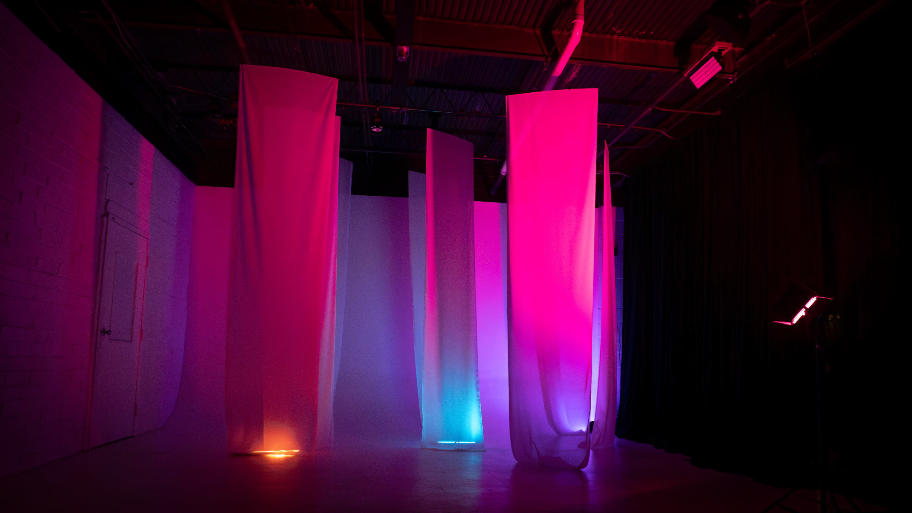 The image shows a studio space with several draped fabrics illuminated by colorful lighting against a plain backdrop.