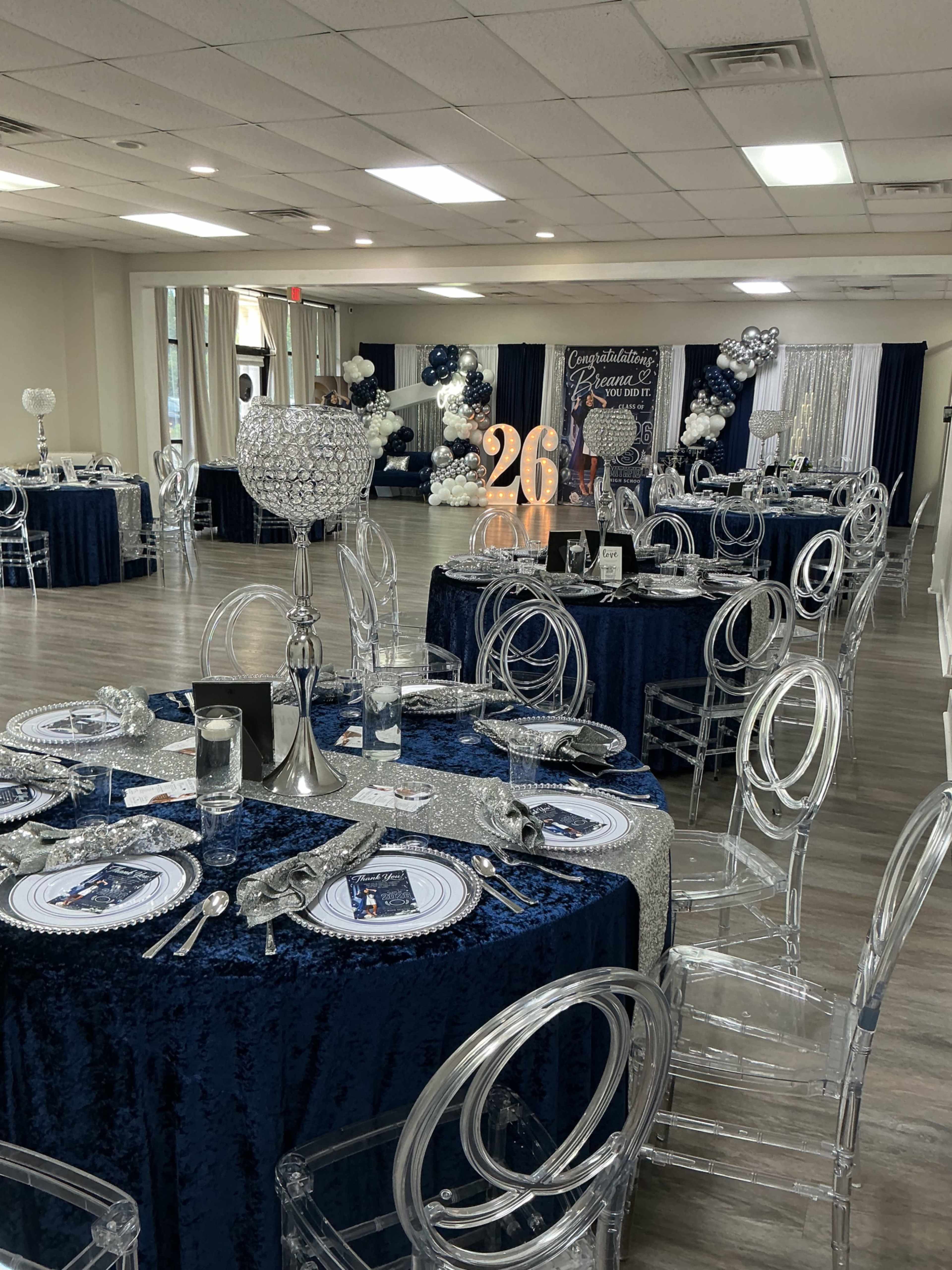 The image shows a decorated event space with round tables covered in navy blue tablecloths, surrounded by clear acrylic chairs, and adorned with elegant centerpieces and balloons.