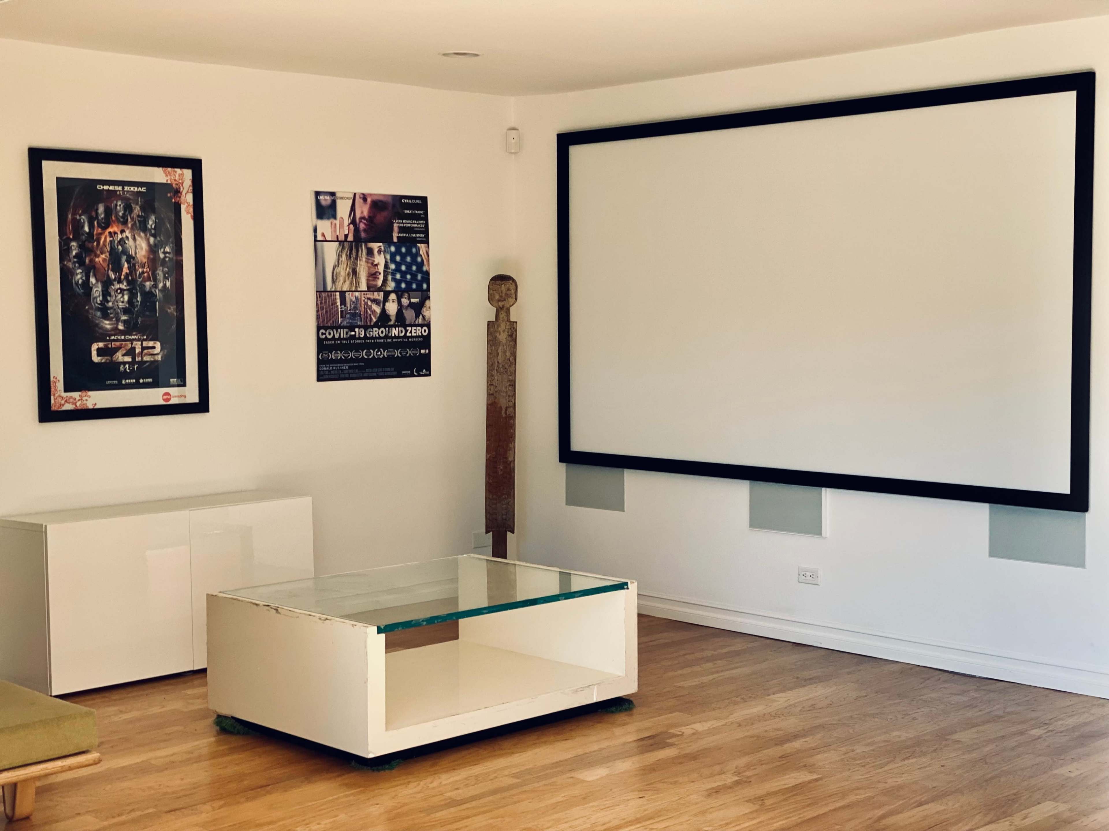 A minimalist room with a large projection screen, a coffee table, and two posters on the wall.