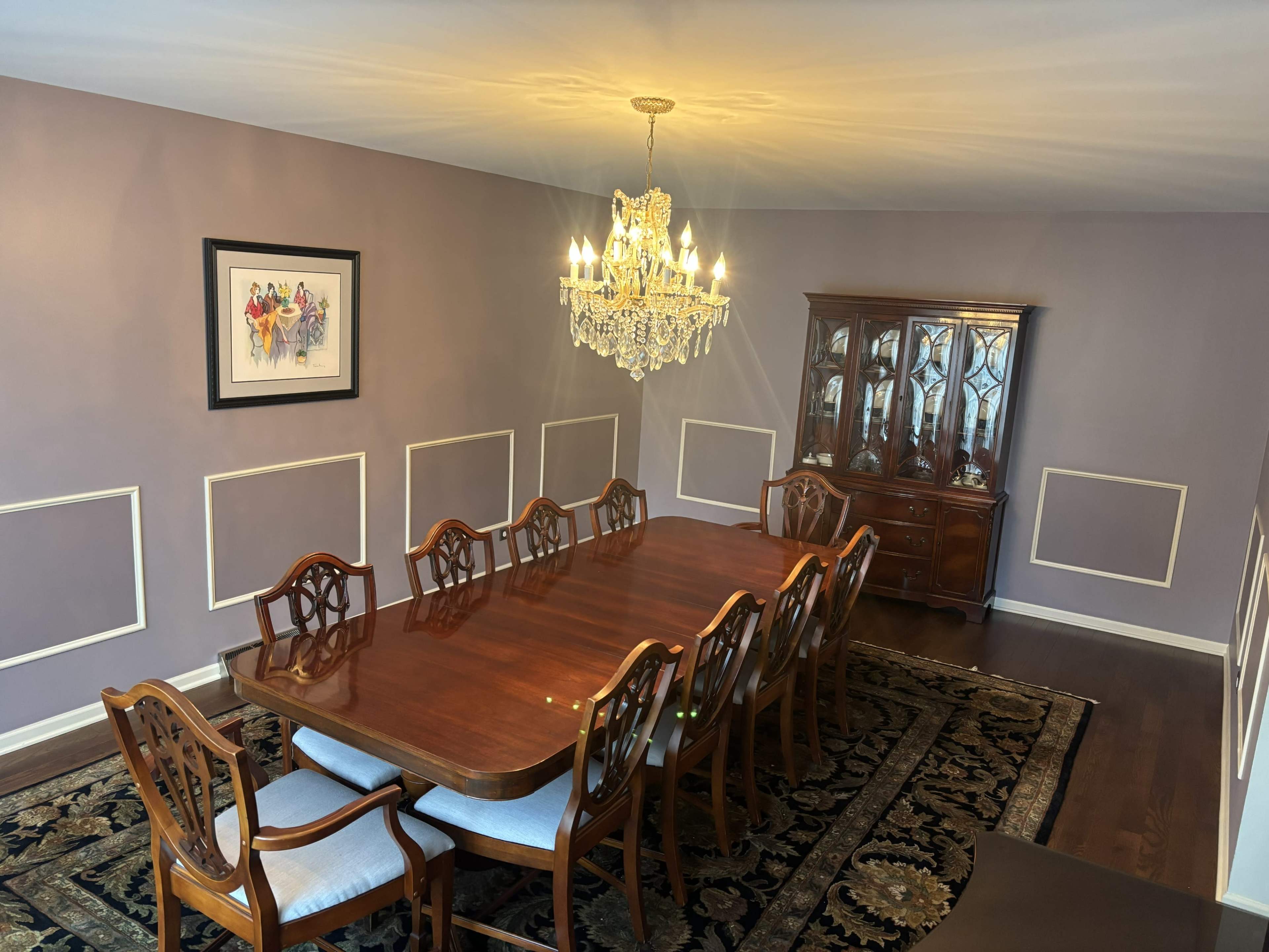 A formal dining room features a long wooden table surrounded by chairs, with a chandelier overhead and a china cabinet against the wall.