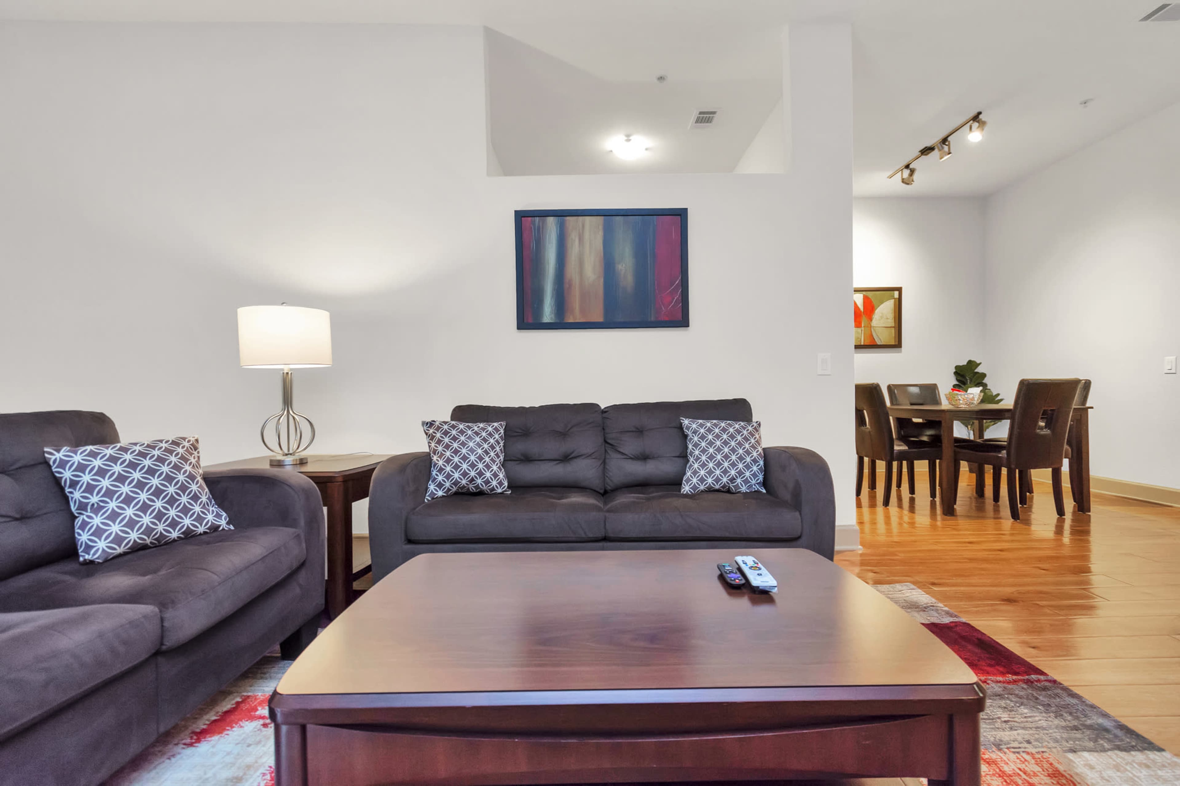 A living room features two dark sofas, a coffee table, and a lamp, with an adjacent dining area visible in the background.