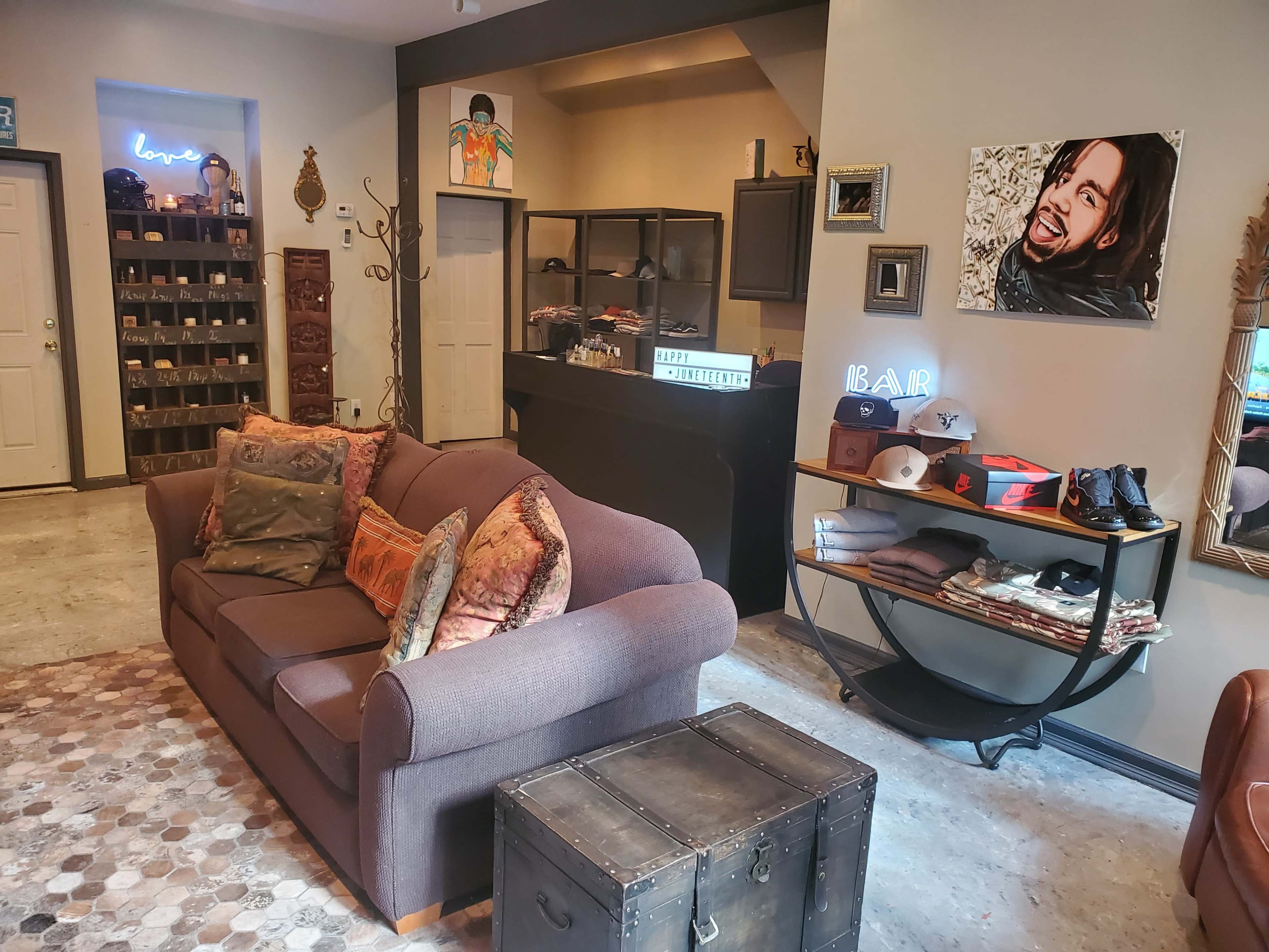 A cozy lounge area featuring a brown sofa, a vintage trunk, and a display shelf filled with hats, alongside a bar counter and various decor on the walls.