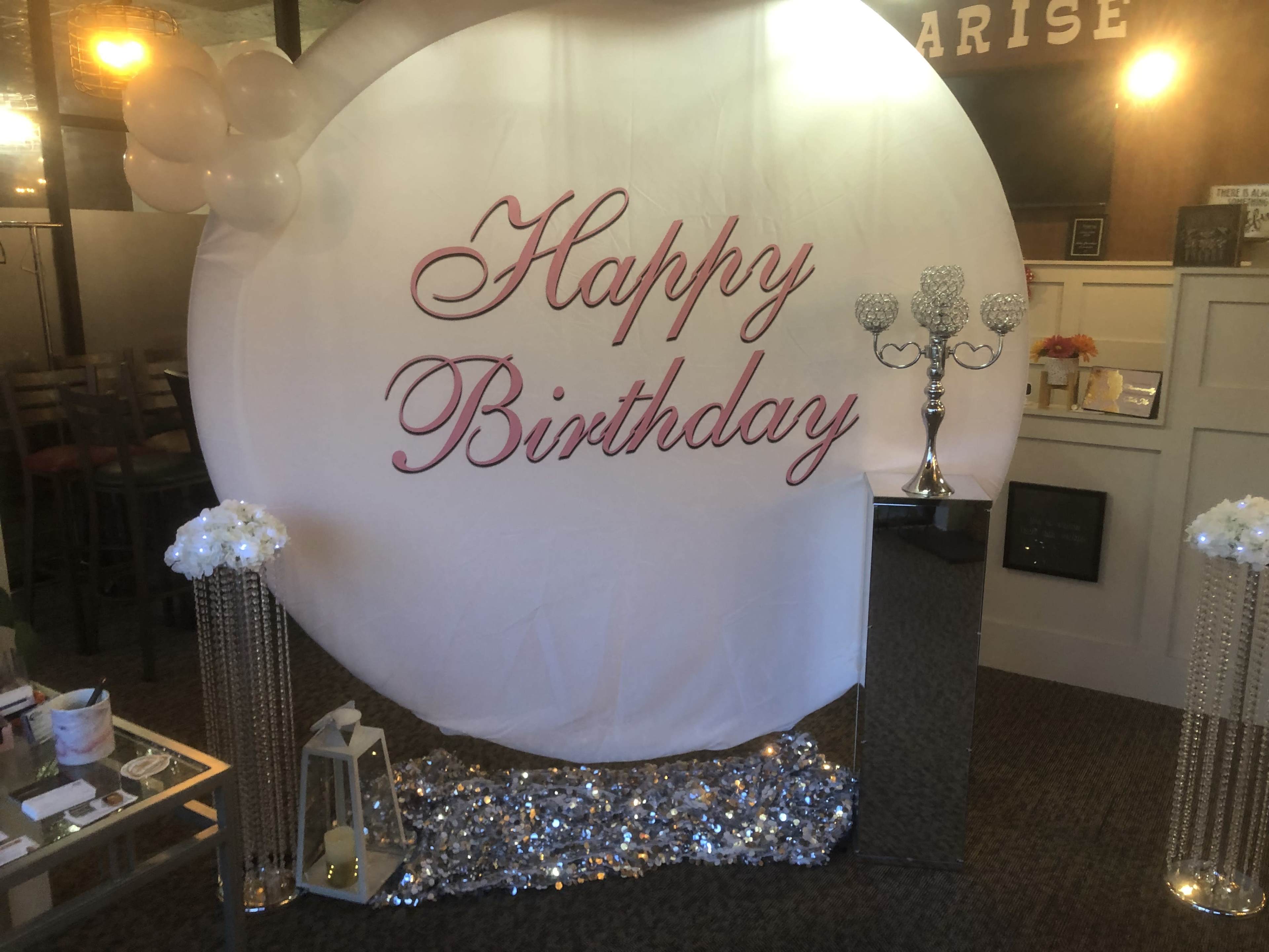 The image shows a decorated space featuring a large circle with the words "Happy Birthday" in script, flanked by floral arrangements and a metallic display.