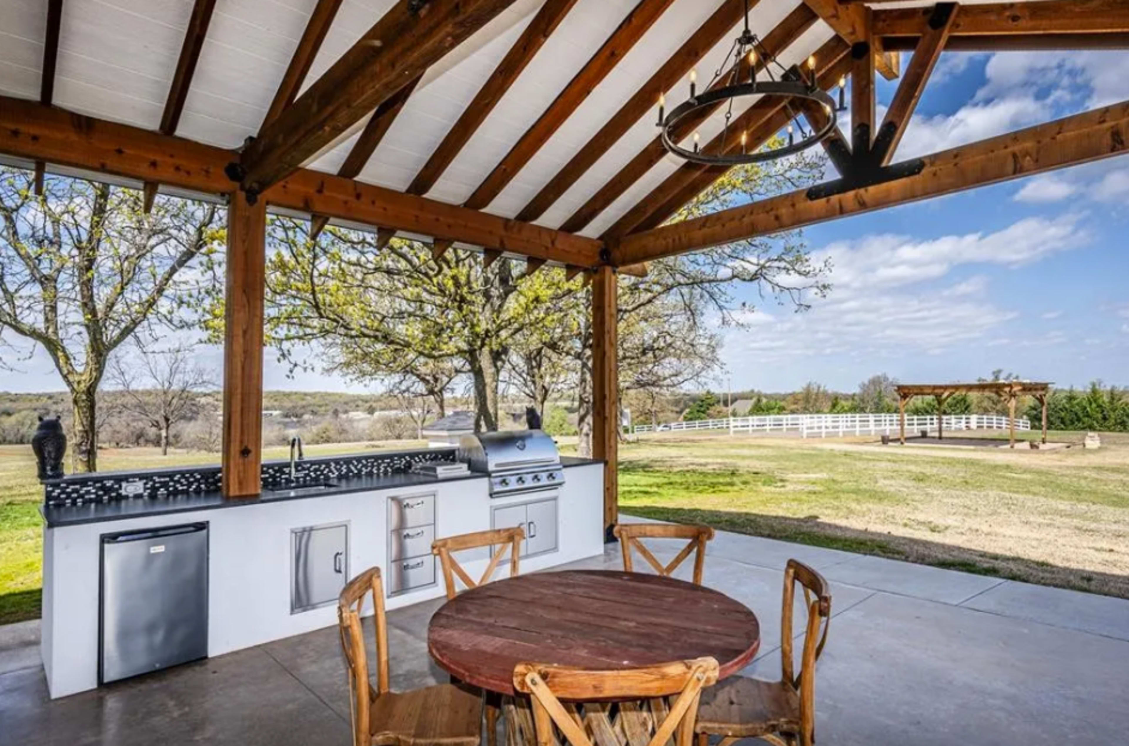 The image shows an outdoor kitchen area with a grill, countertop, and a wooden dining table beneath a covered structure, surrounded by a grassy landscape and trees.