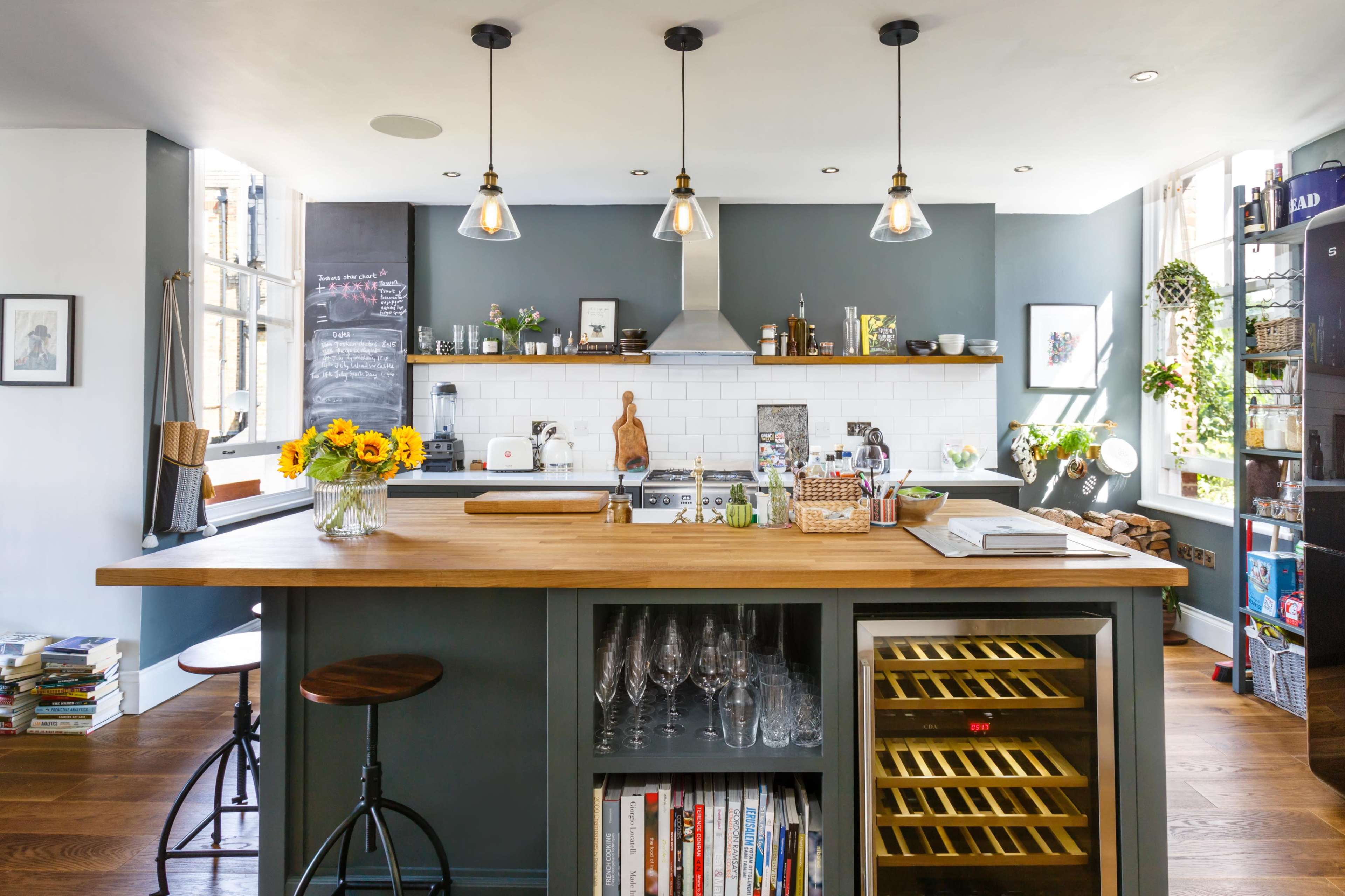 A modern kitchen features a central wooden island with bar stools, a wine fridge, and shelves filled with glassware and cookbooks.