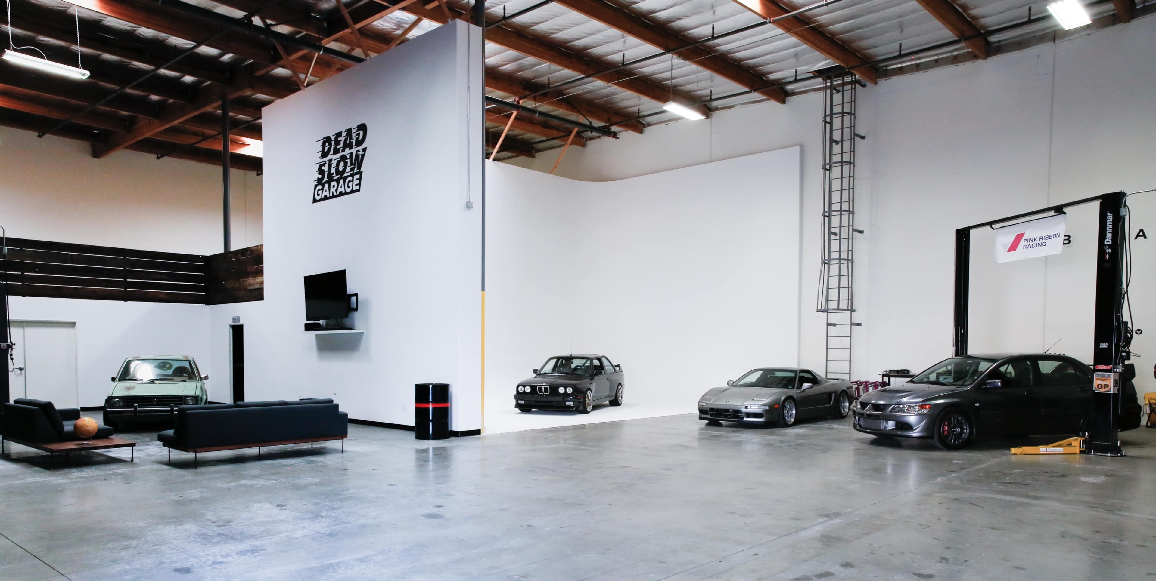 A spacious garage featuring several classic cars, a large white wall, and minimal furnishings, with the words "DEAD SLOW GARAGE" displayed on the wall.