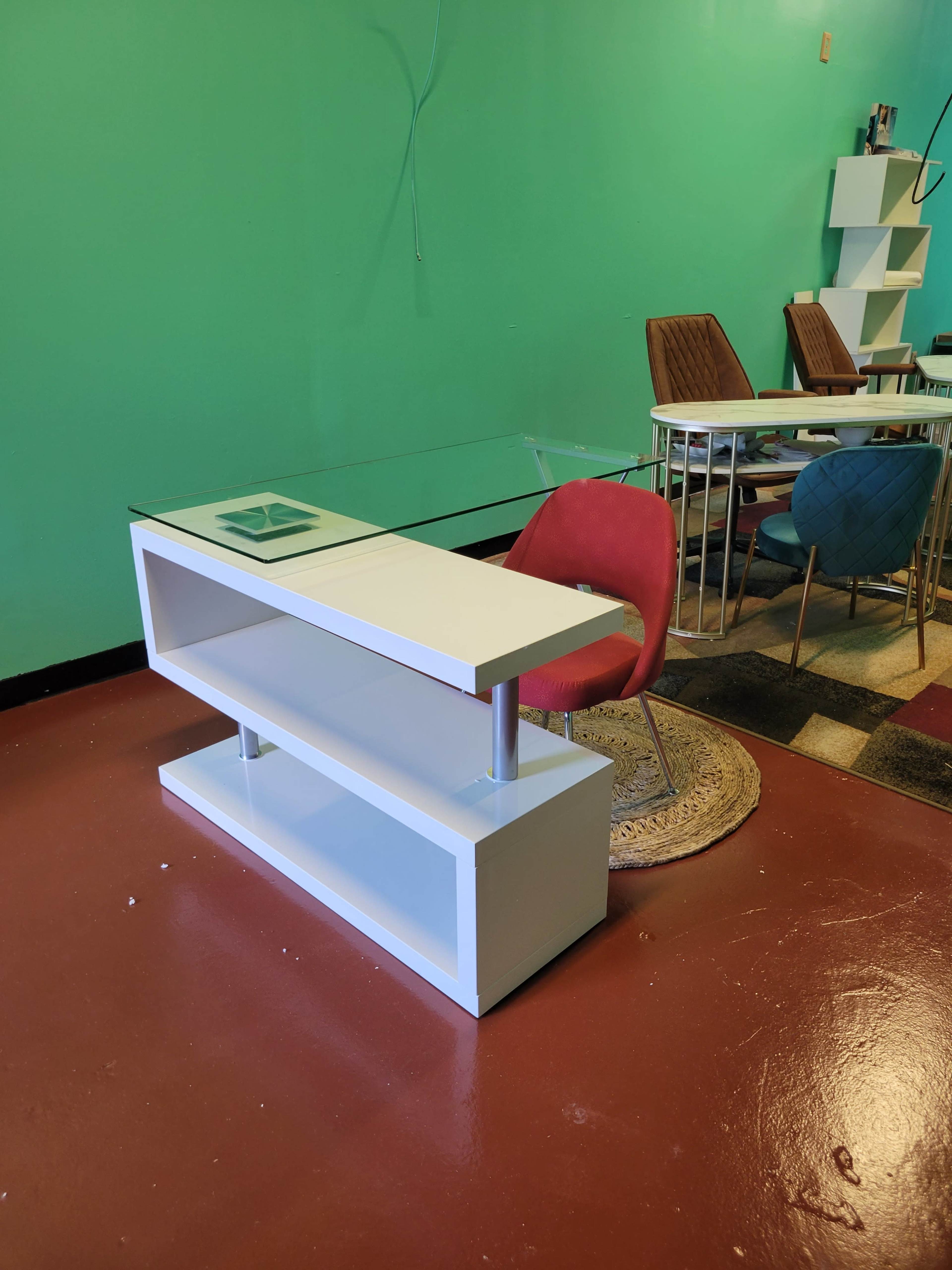 A modern glass-top desk with a white base is positioned in a room featuring a green wall and various chairs.