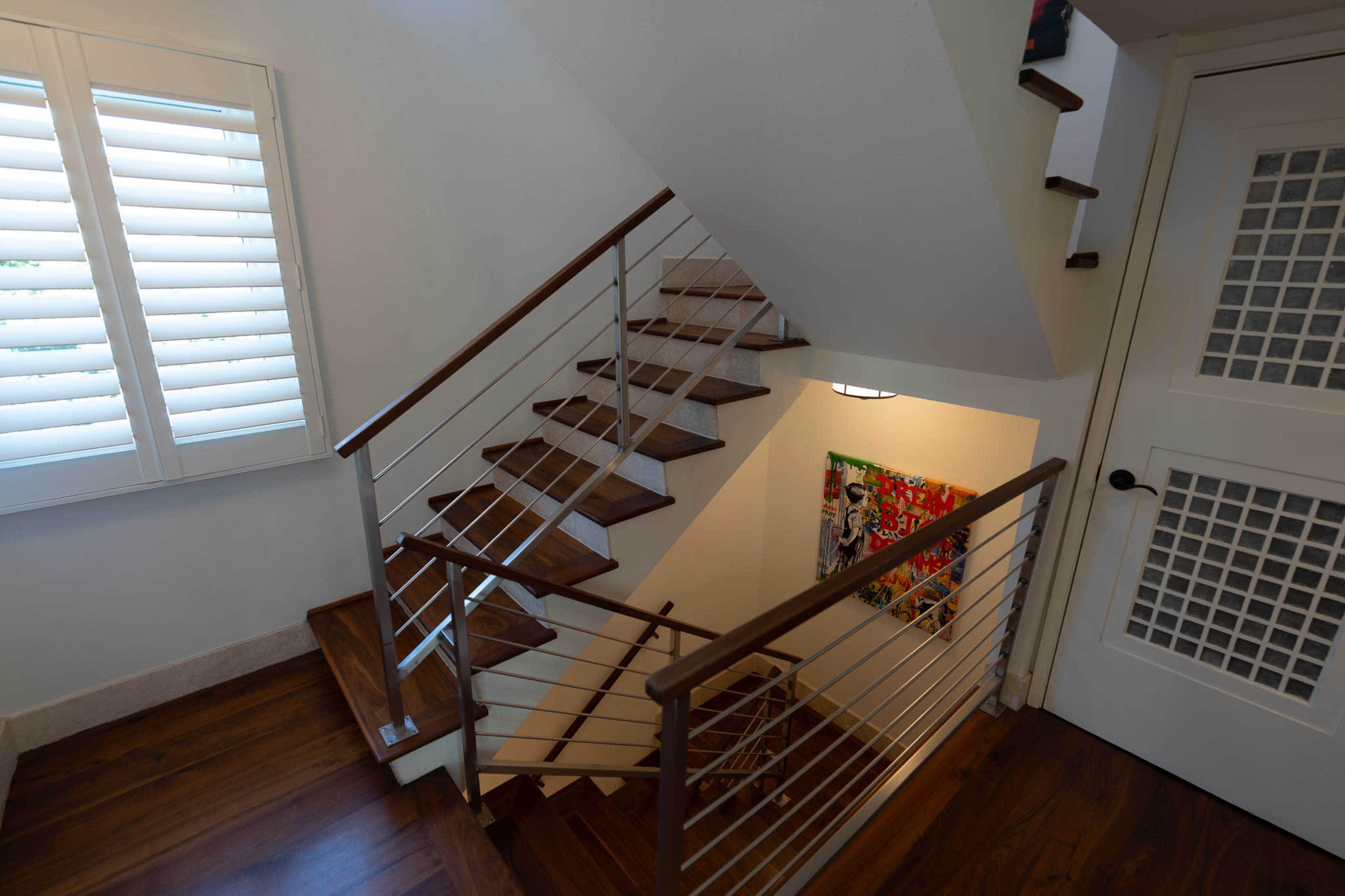 The image shows a modern staircase with wooden steps and a metal railing, leading down to a landing with a colorful artwork on the wall.