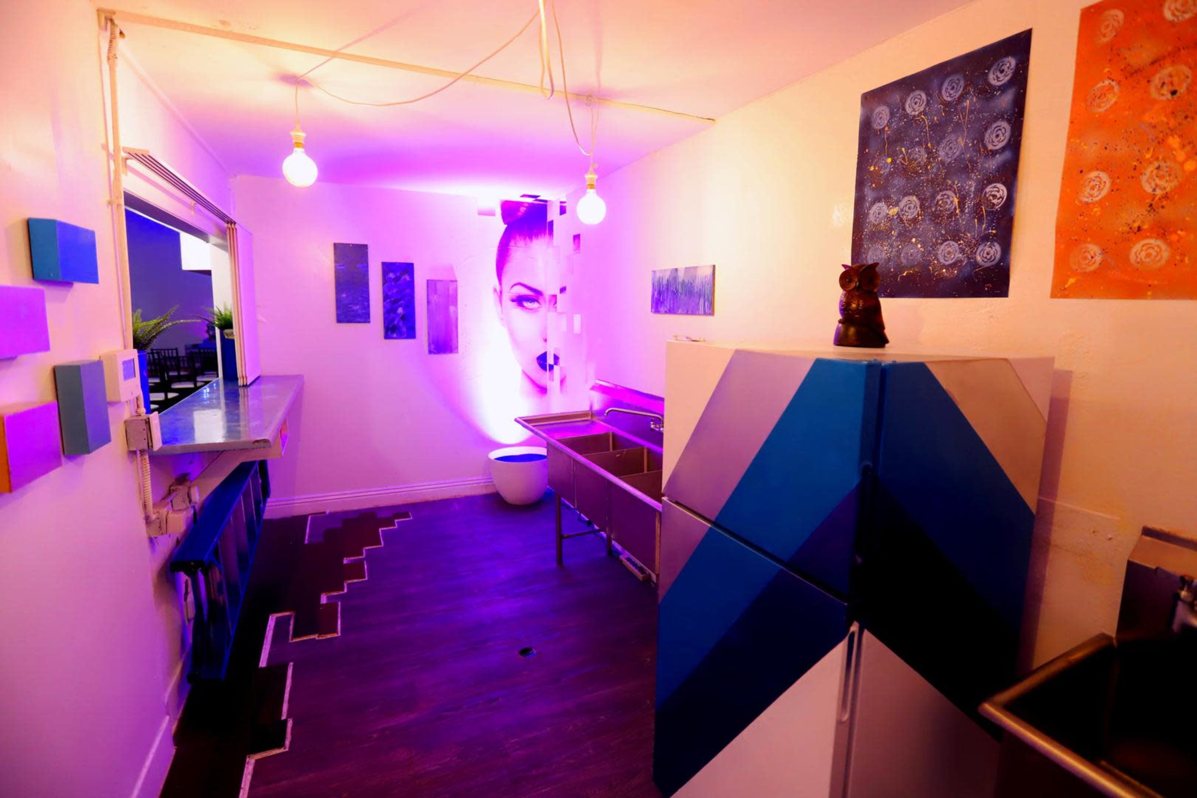 The image shows a brightly lit room featuring a colorful wall, modern art pieces, a blue and white striped refrigerator, and minimalist furniture.