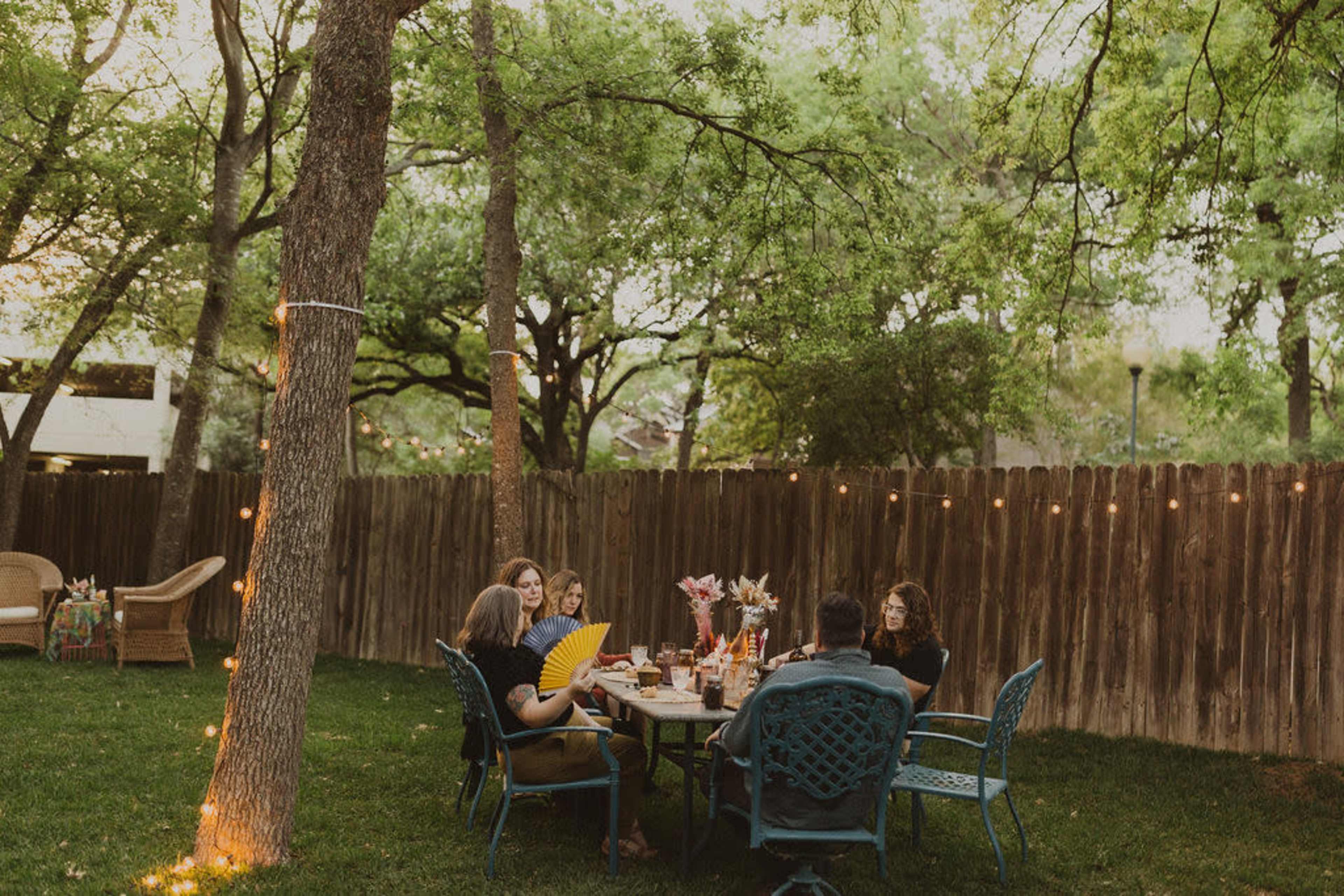 A group of five people is seated around a table in a backyard, with string lights illuminating the scene and trees surrounding the area.