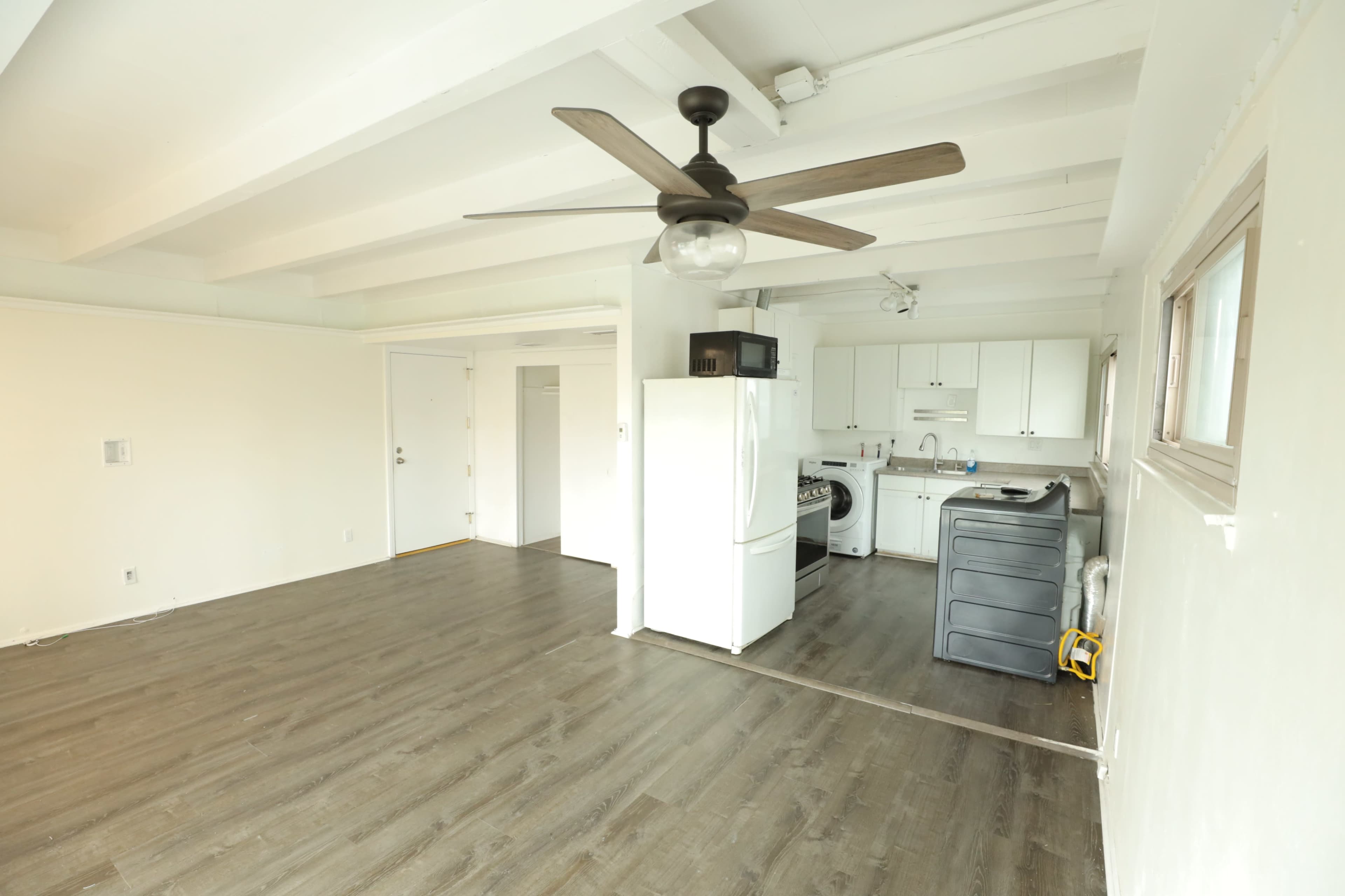 The image shows an empty room with a ceiling fan, a white refrigerator, a stove, and a washing machine, featuring wooden flooring and white walls.