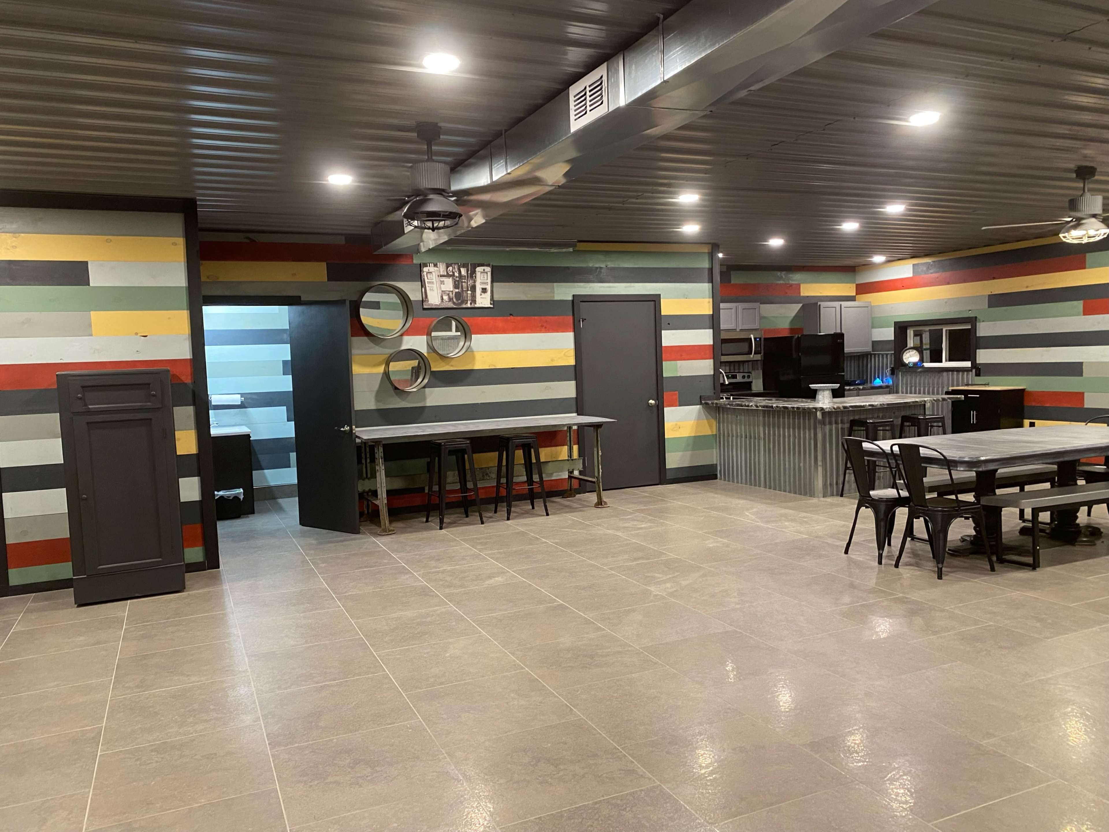 The image shows a spacious room with multicolored striped walls, a kitchen area, and dining tables arranged in the center.