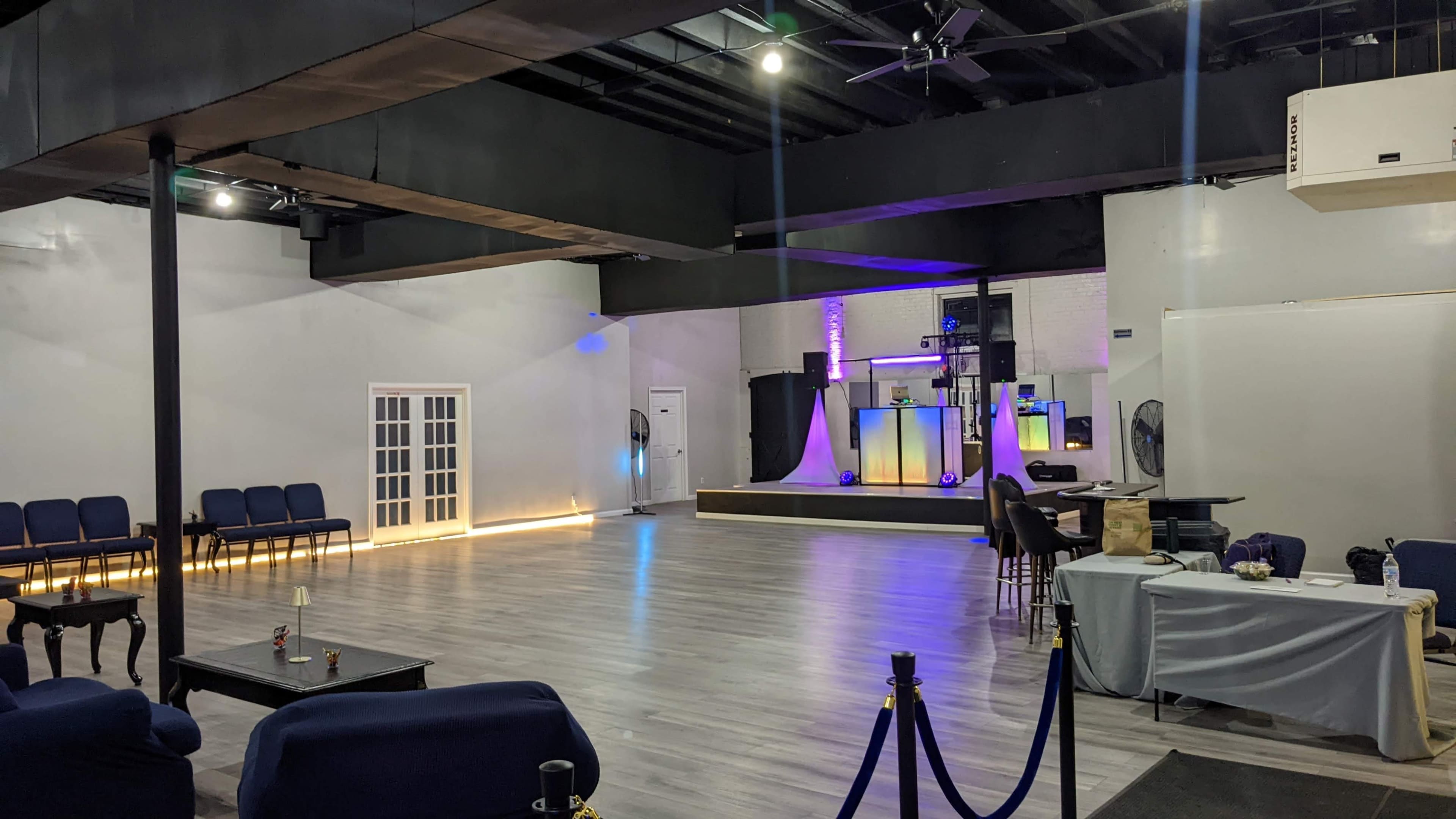 A spacious indoor venue with a wooden floor, seating areas, and a raised stage featuring colorful lighting.