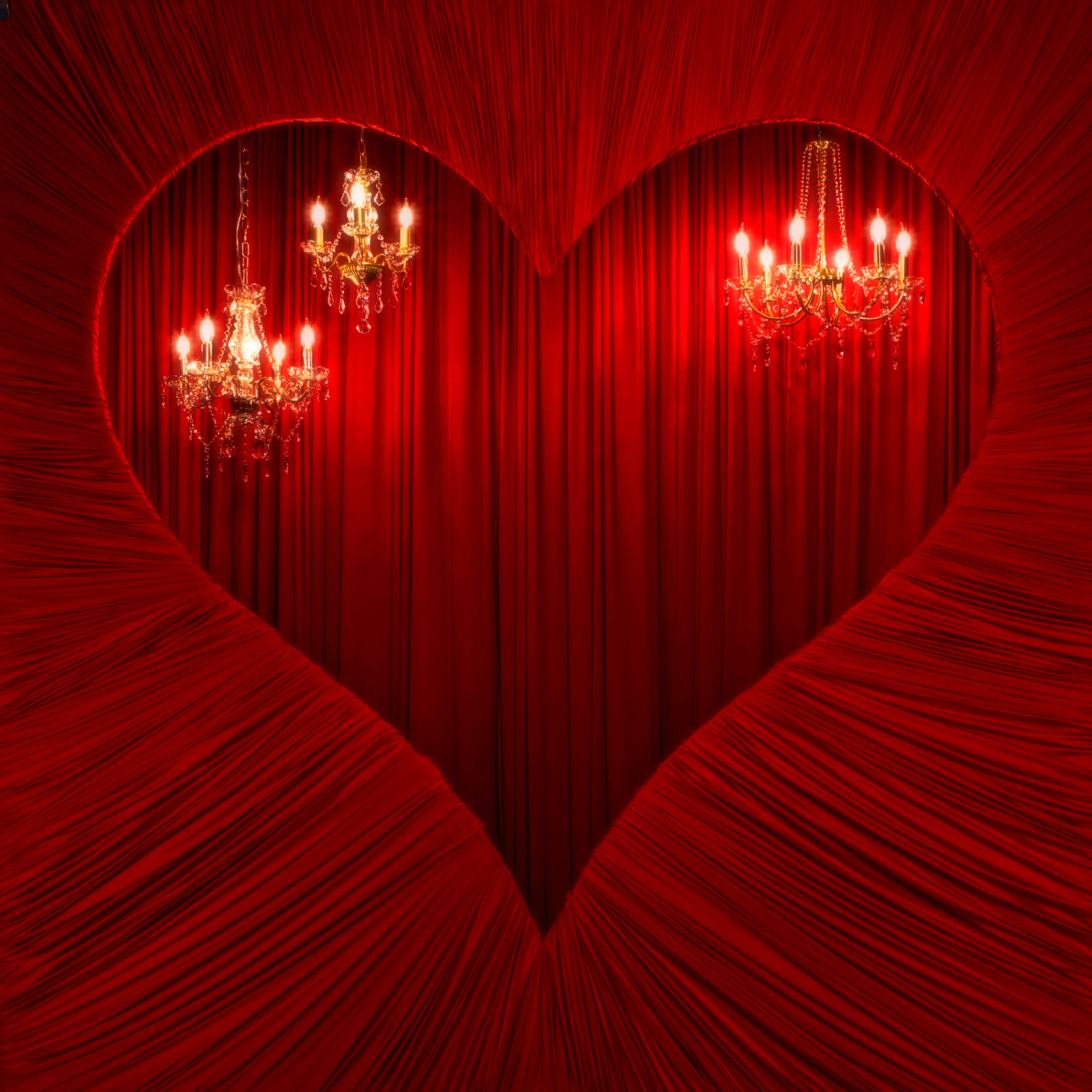 The image features a large heart-shaped opening in red draped fabric, revealing three ornate chandeliers suspended against a similarly red backdrop.