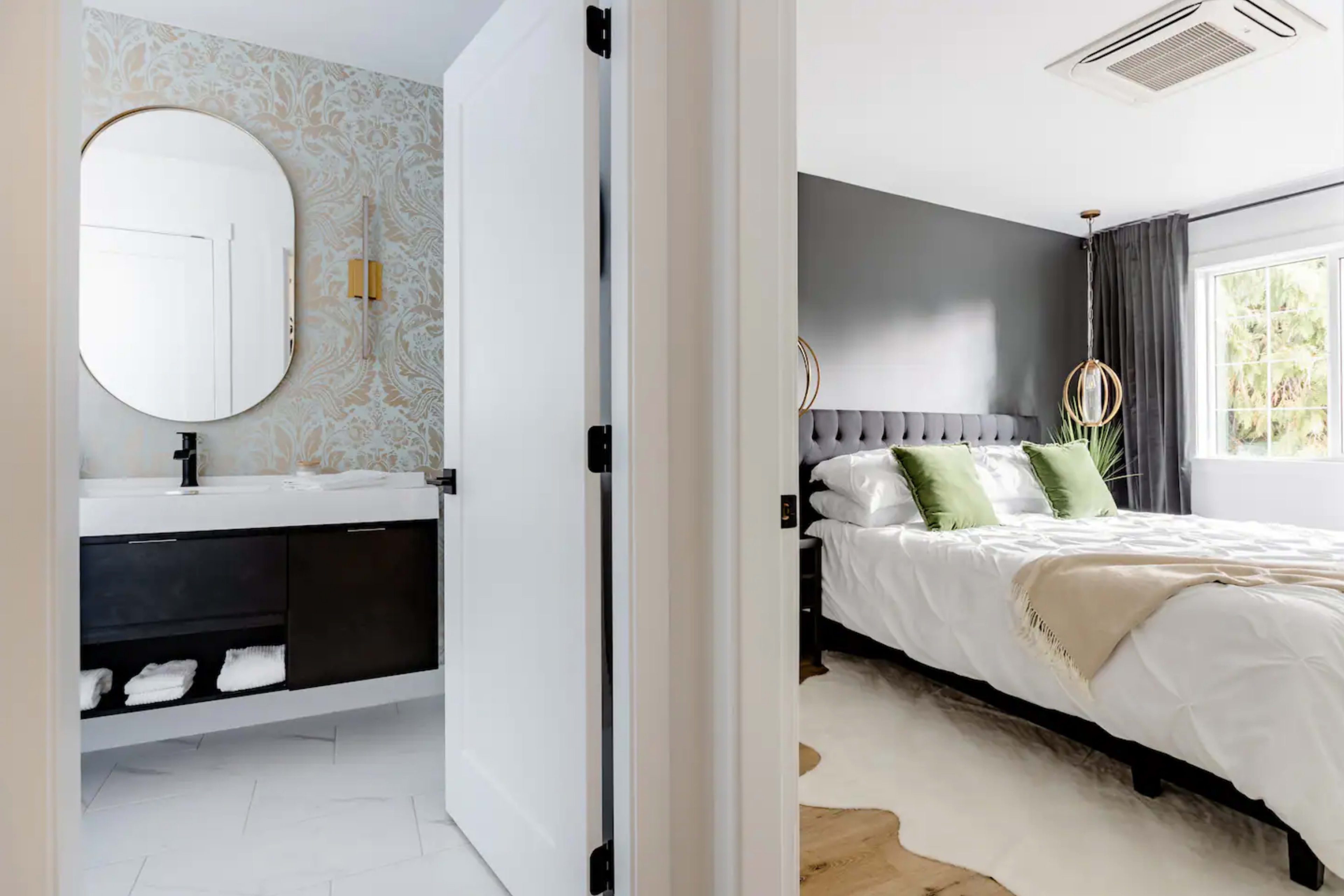 A doorway leads from a modern bathroom with a patterned wall to a bedroom featuring a bed with green accent pillows and large windows.