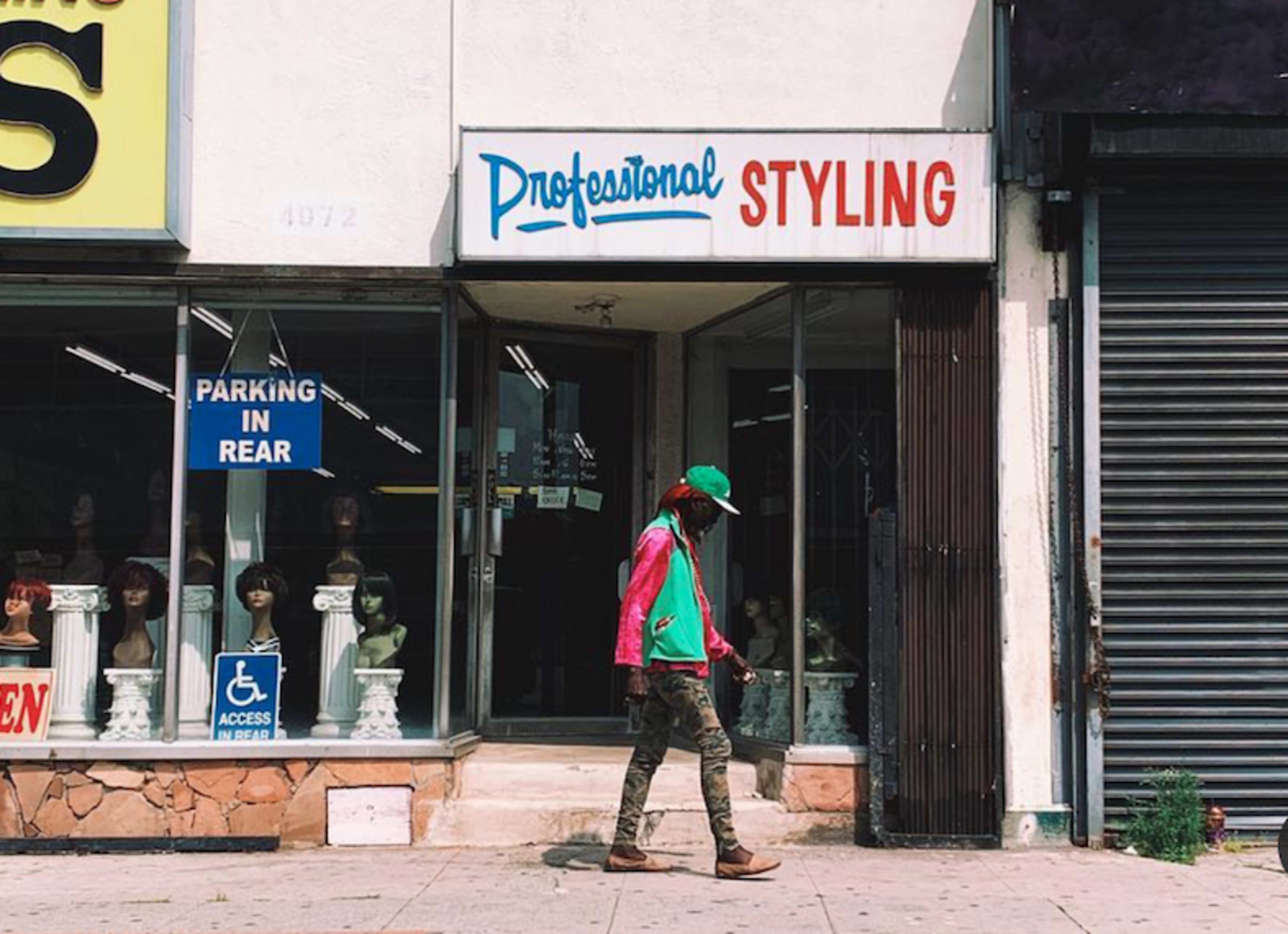 A person wearing a colorful jacket and cap walks past a storefront advertising "Professional Styling," featuring mannequins with wigs inside.