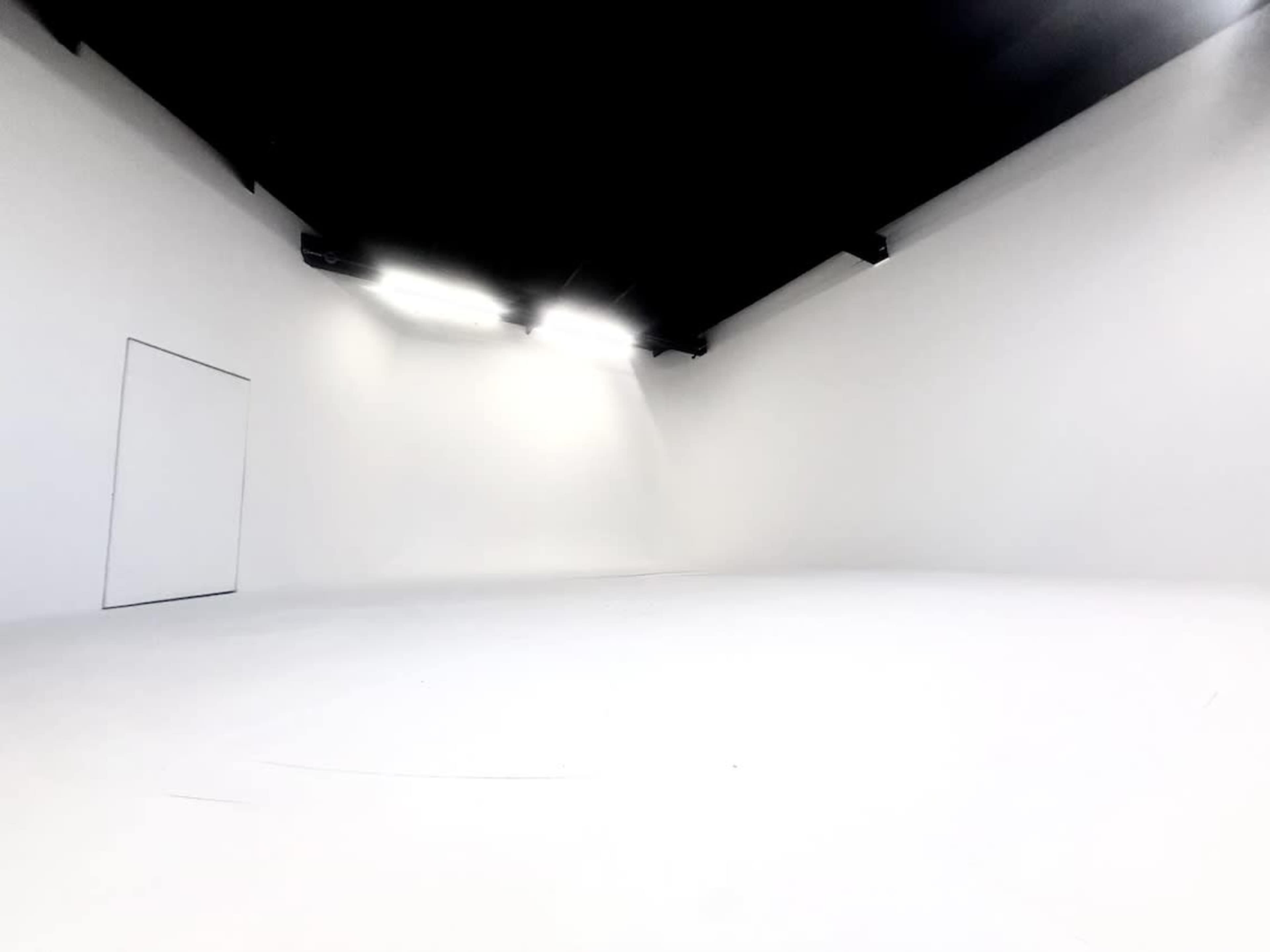 The image shows a spacious, empty room with white walls and a white floor, illuminated by overhead lights.