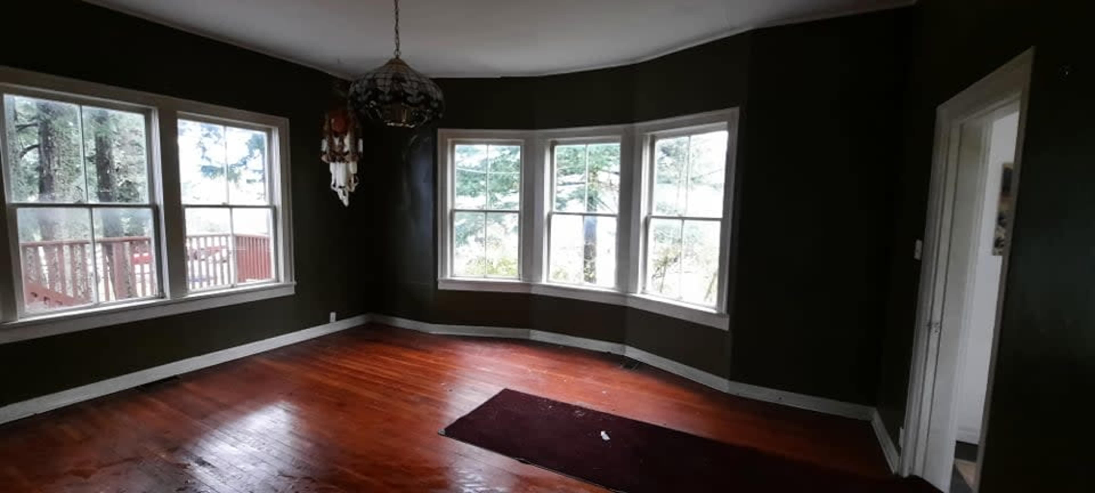 The image shows an empty room with dark green walls, large windows letting in natural light, and hardwood floors.