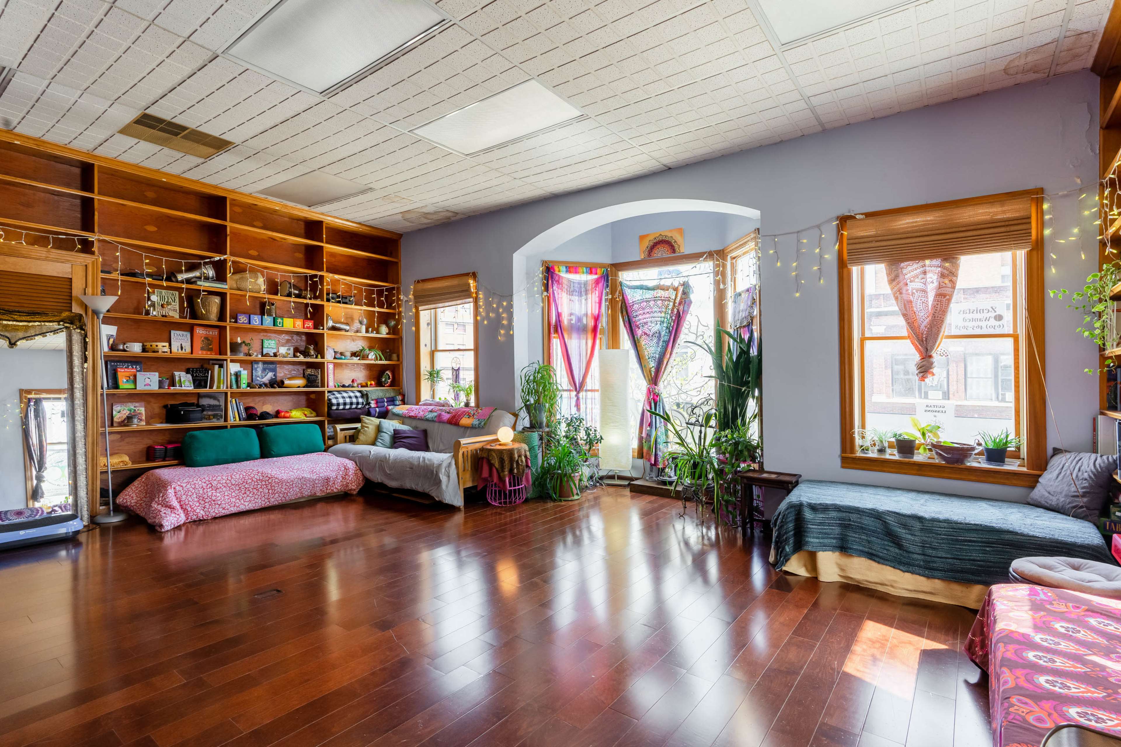 A spacious, brightly lit room with hardwood floors, large windows, and shelves filled with decorative items and plants.