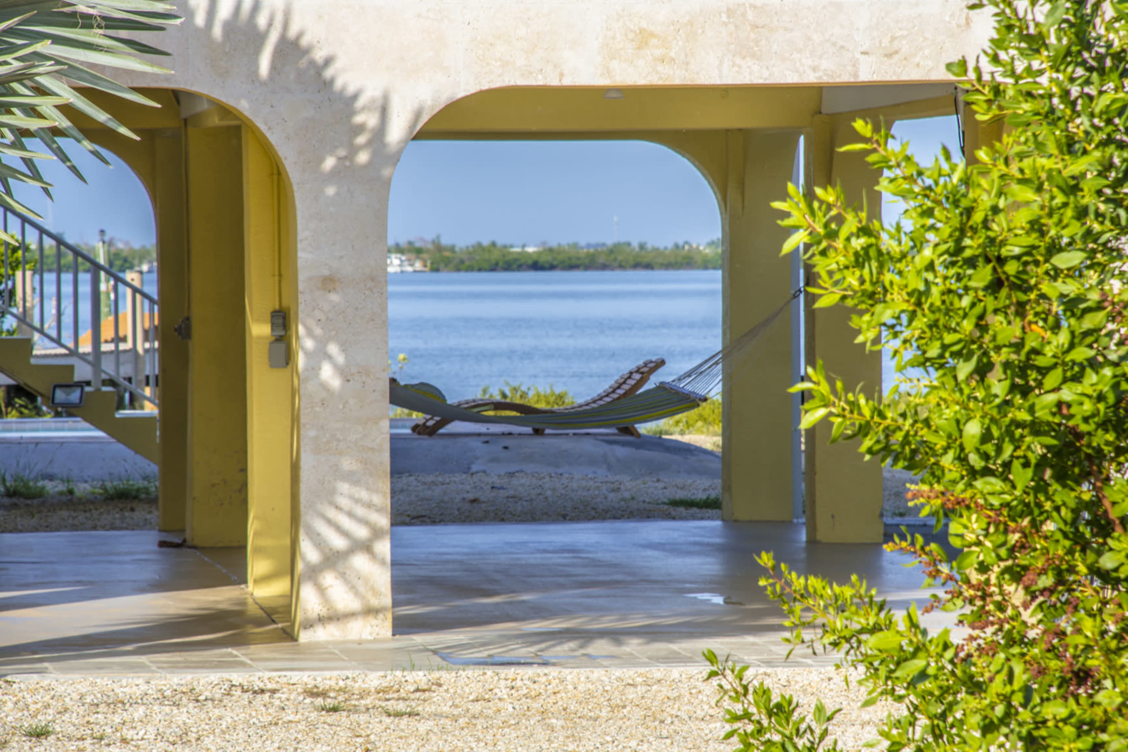 A hammock is suspended between two posts, framed by arches of a building, with a view of a calm body of water in the background.