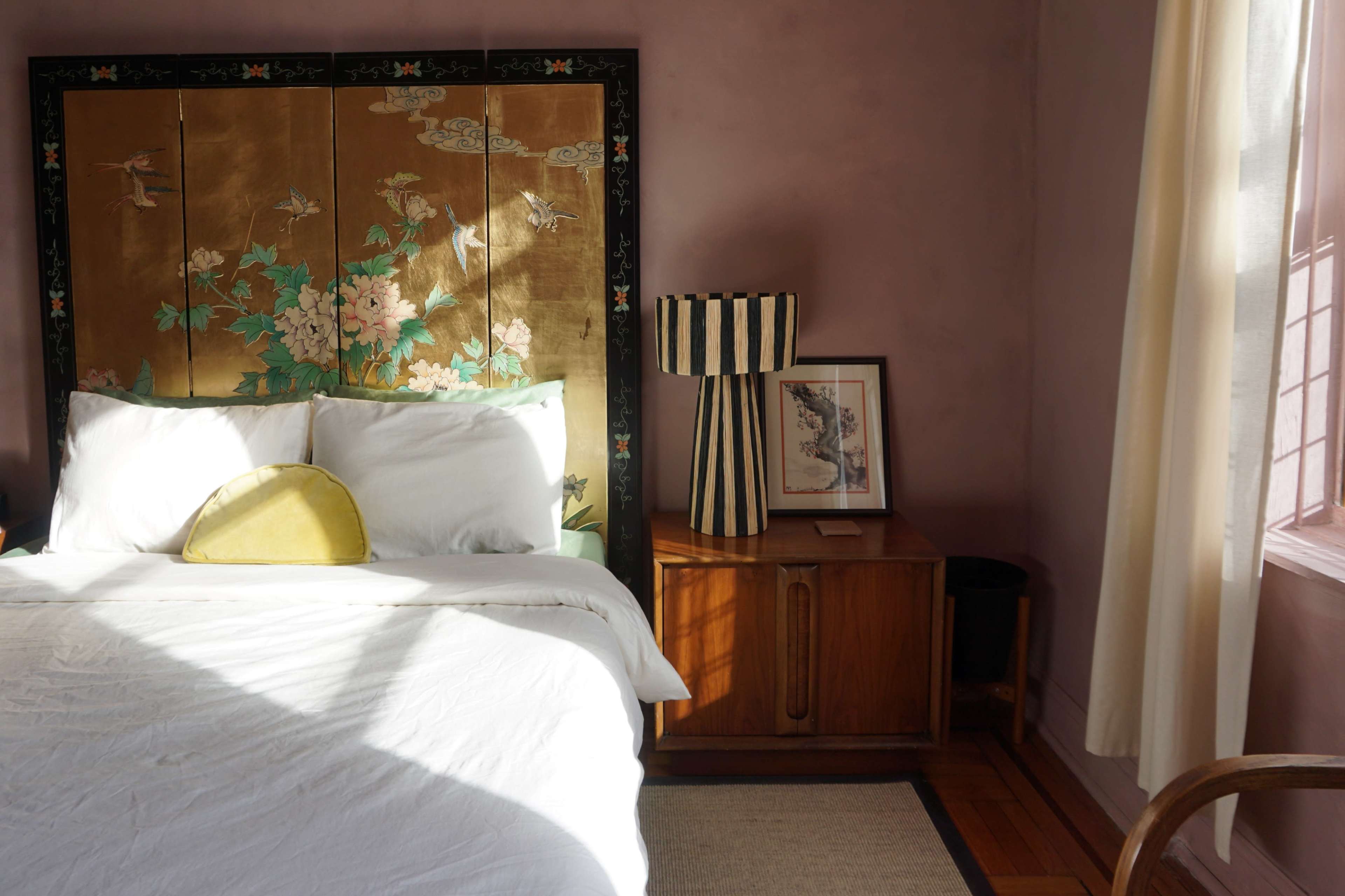 A bedroom with a bed featuring white linens, a decorative headboard with floral designs, a striped lamp, and a wooden cabinet beside it.