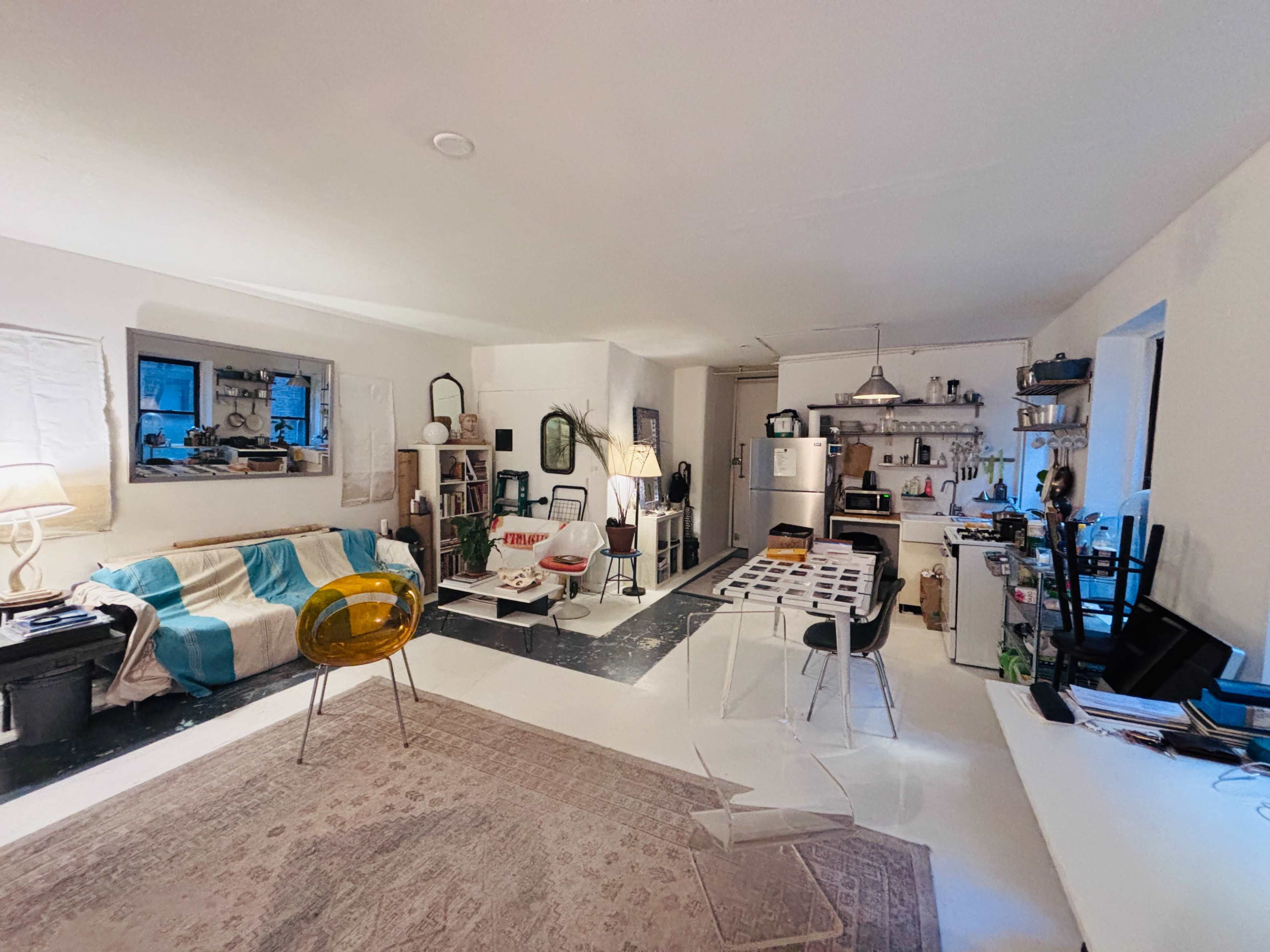 The image shows a modern, open-plan living space with a living area featuring a couch, chairs, and a rug, leading to a kitchen with various appliances and furnishings.