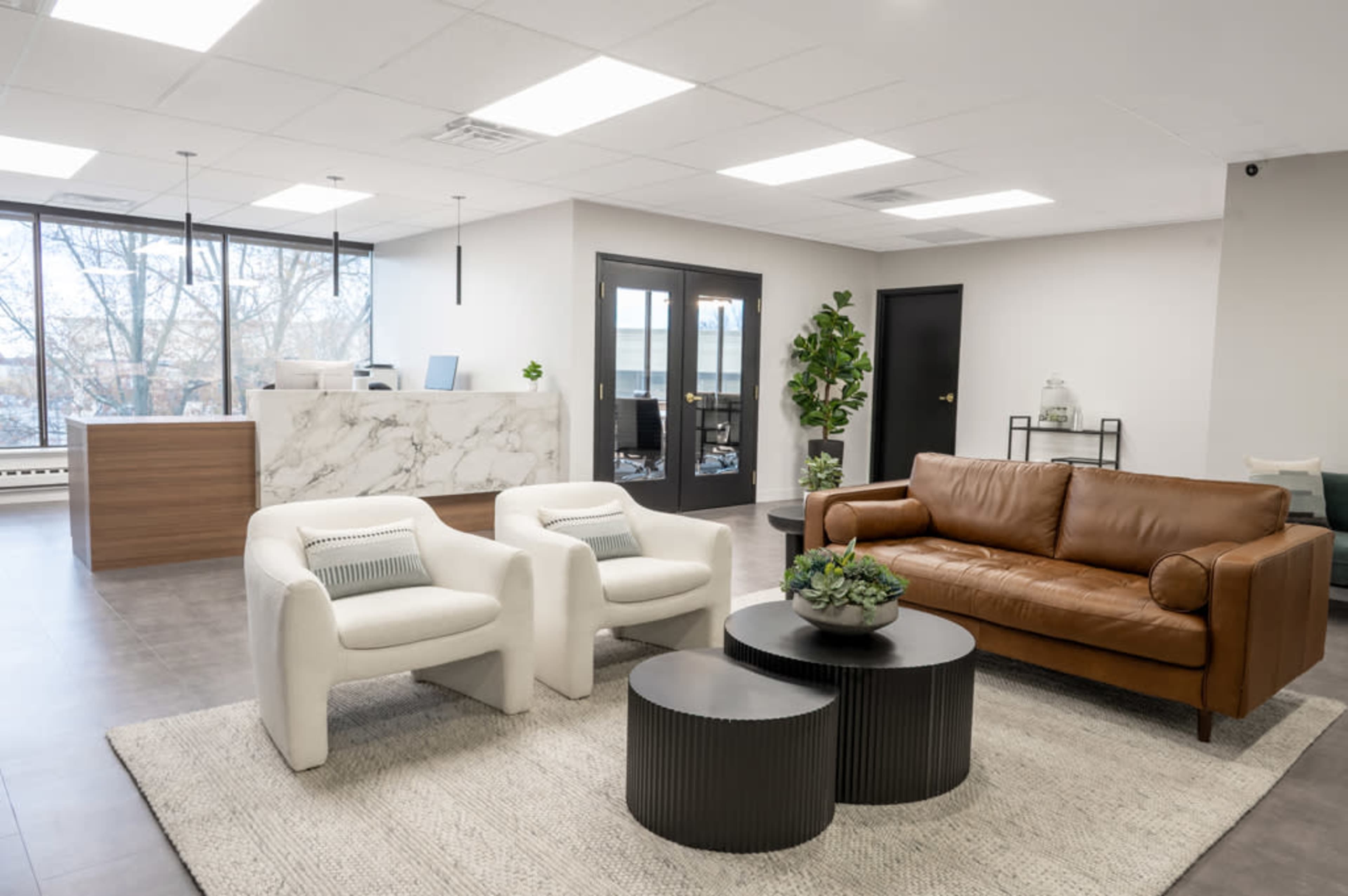 The image shows a modern office reception area with a marble front desk, two white armchairs, a brown leather sofa, and two black coffee tables on a light rug.