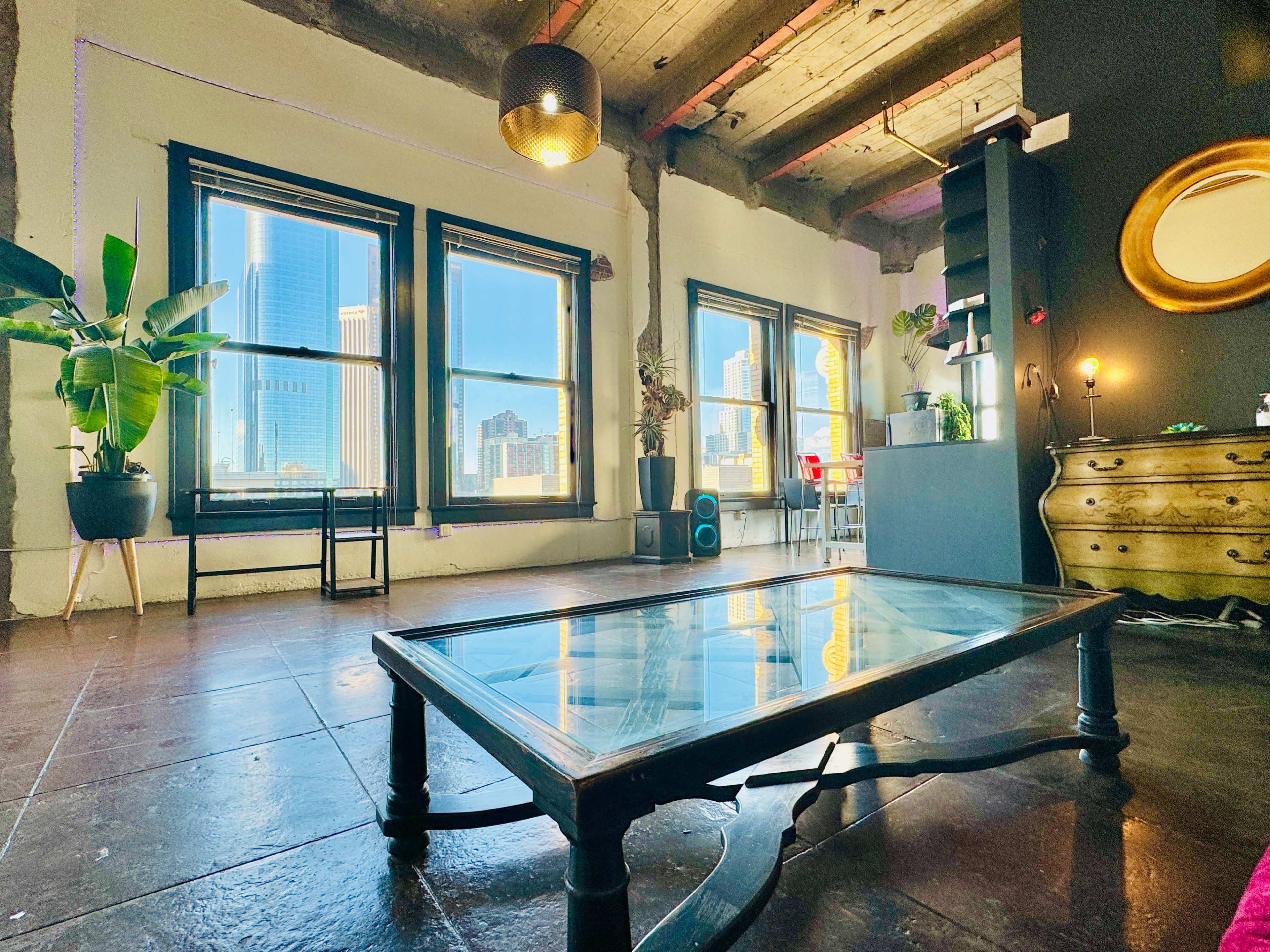 A modern interior with a glass-top coffee table, large windows offering views of a city skyline, and various indoor plants.