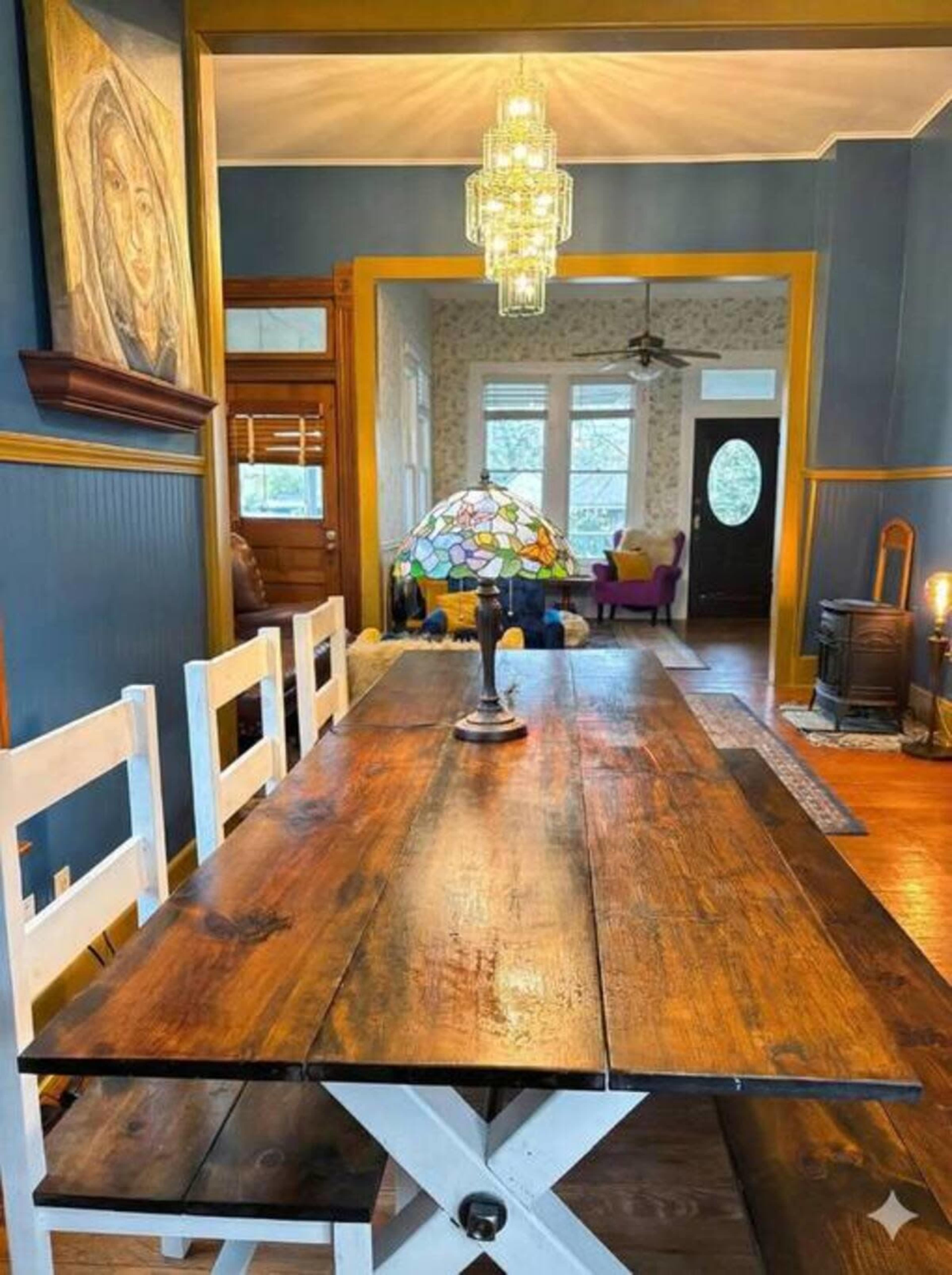 A long wooden dining table sits in a well-lit room featuring blue walls, a stained glass lamp, and a doorway leading to another space with colorful furniture.