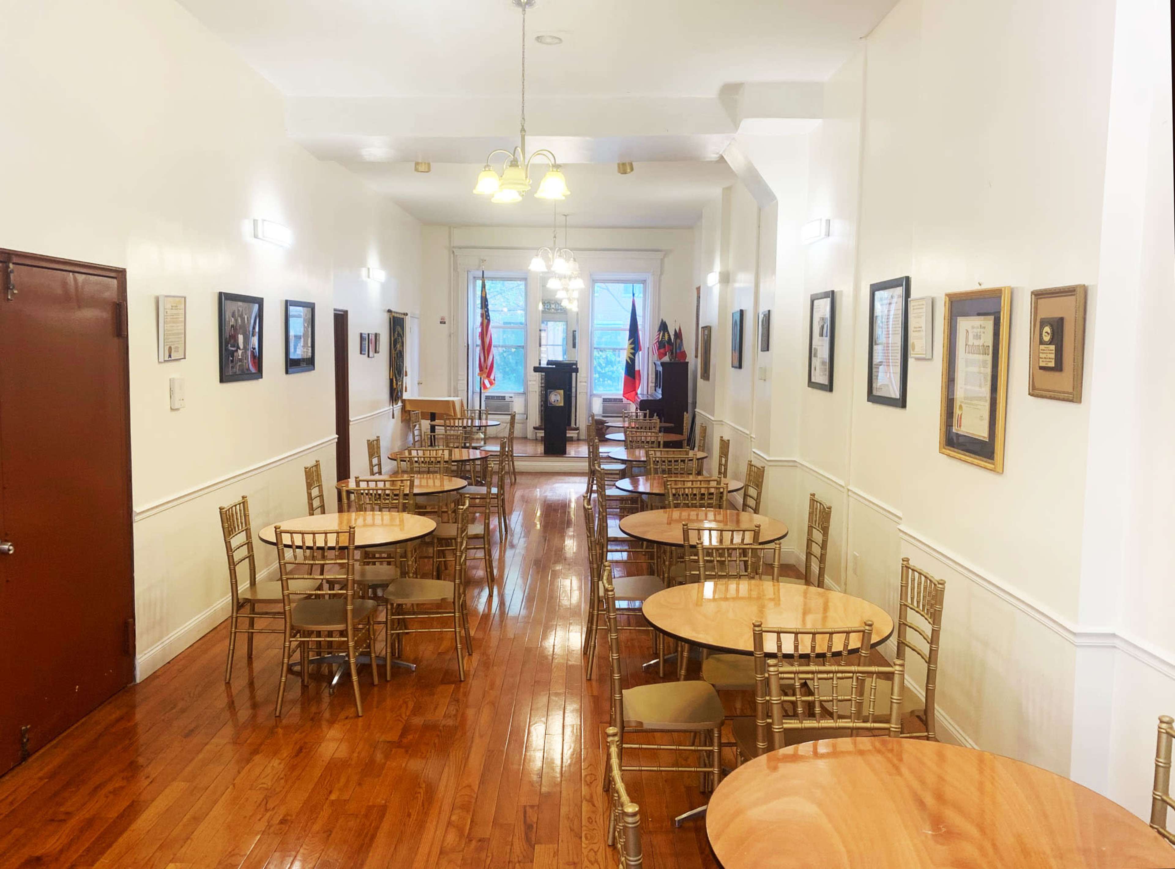 The image shows a well-lit dining hall with wooden floors, several round tables arranged with chairs, and framed documents and photos on the walls.