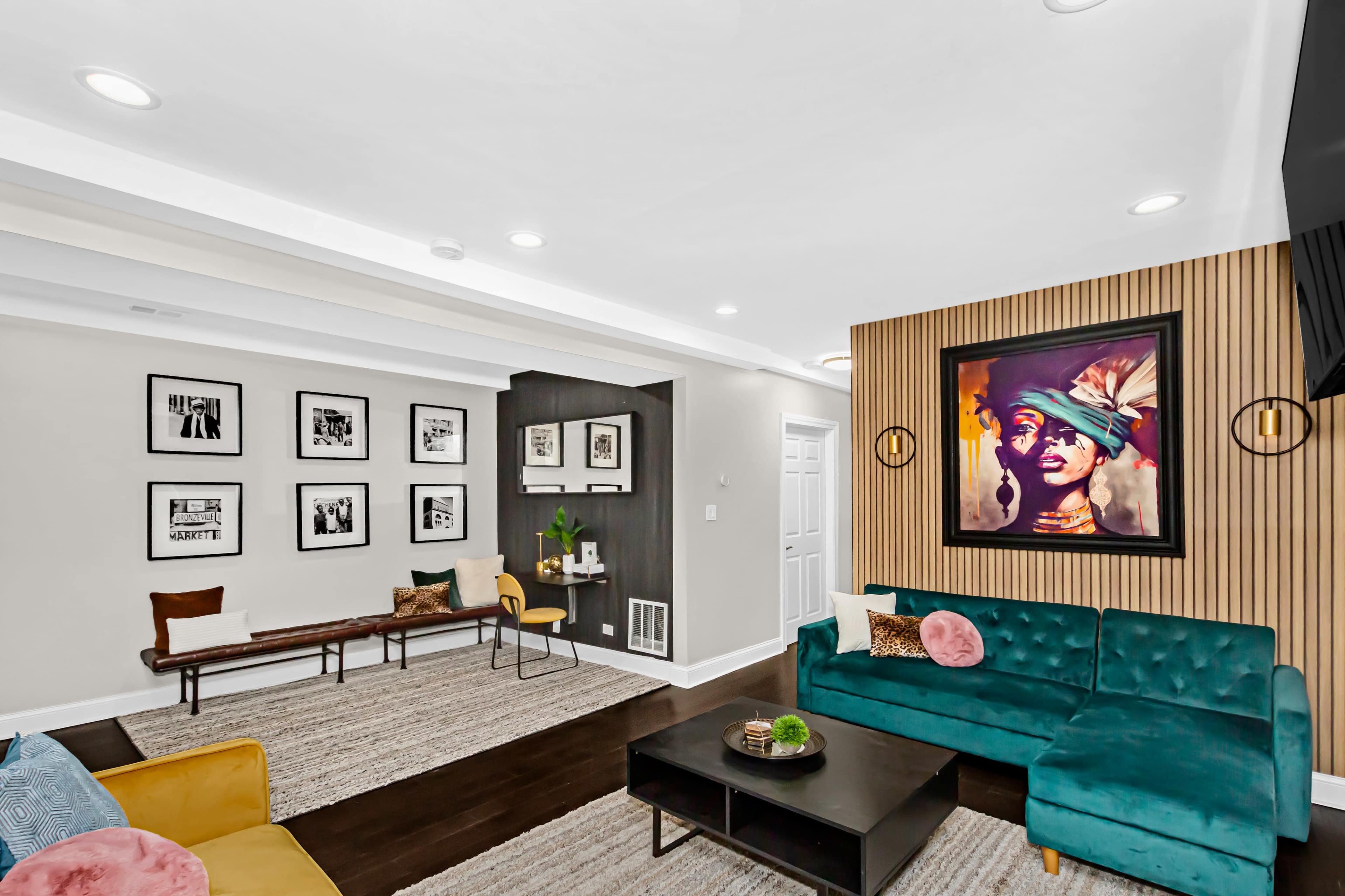 A modern living room with a teal sectional sofa, a wooden bench for seating, framed black-and-white photographs on the walls, and a large colorful artwork above the couch.
