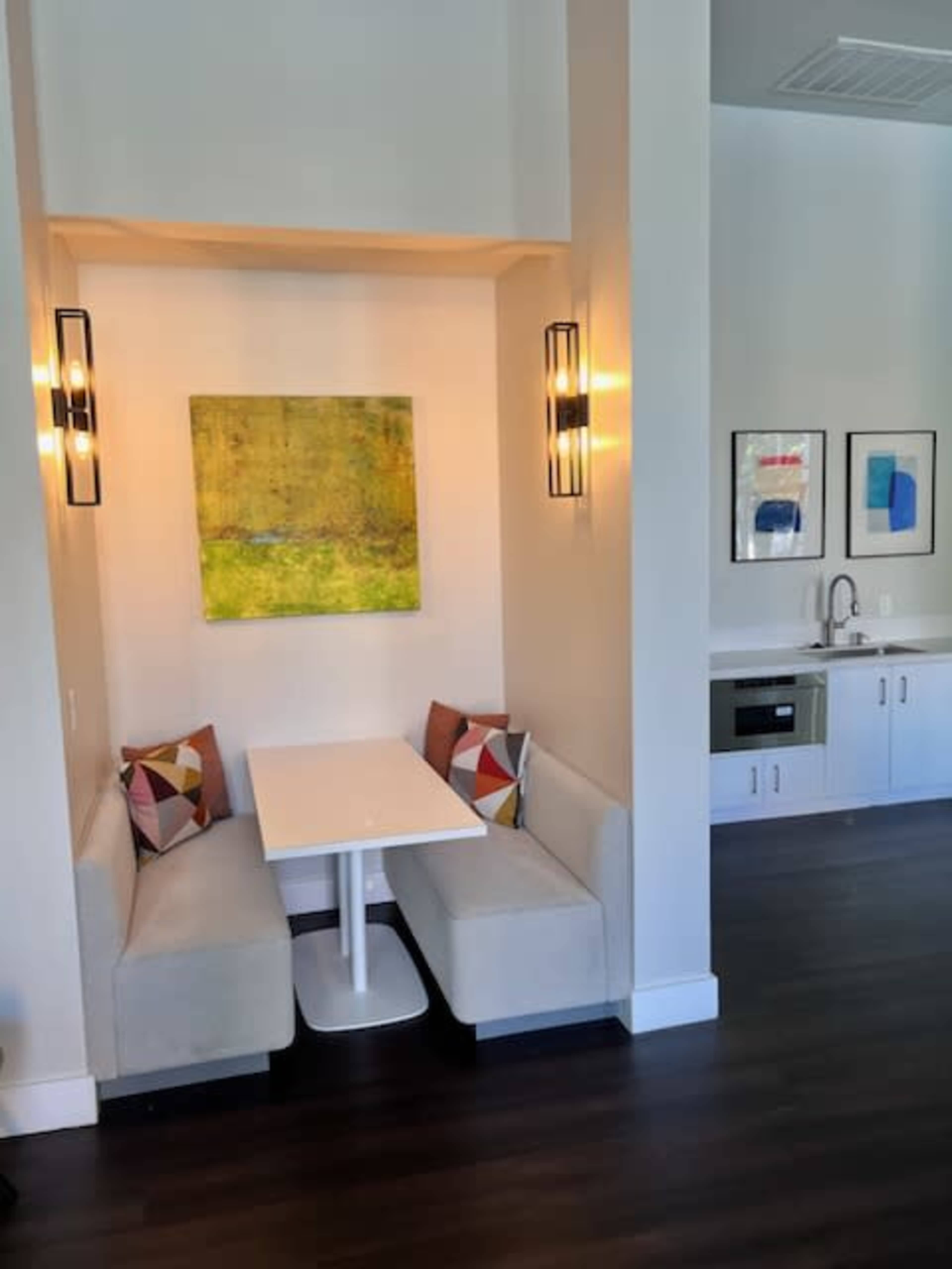 A small dining area features a white table and cushioned seating against a wall, with a green artwork and framed prints nearby.