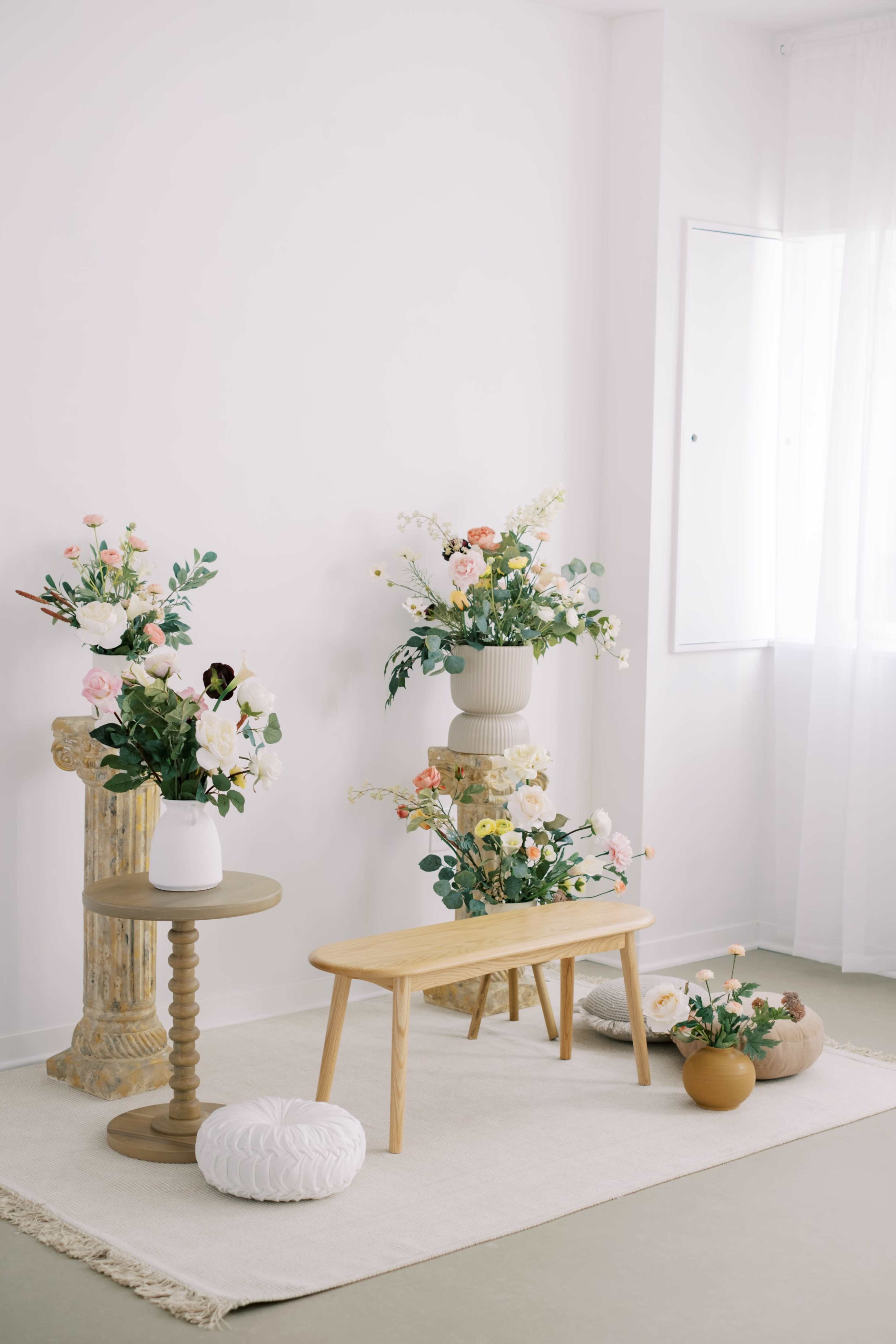 The image features a minimalist interior space with a wooden bench and tables adorned with various flower arrangements in decorative vases, set on a light rug against a white wall.