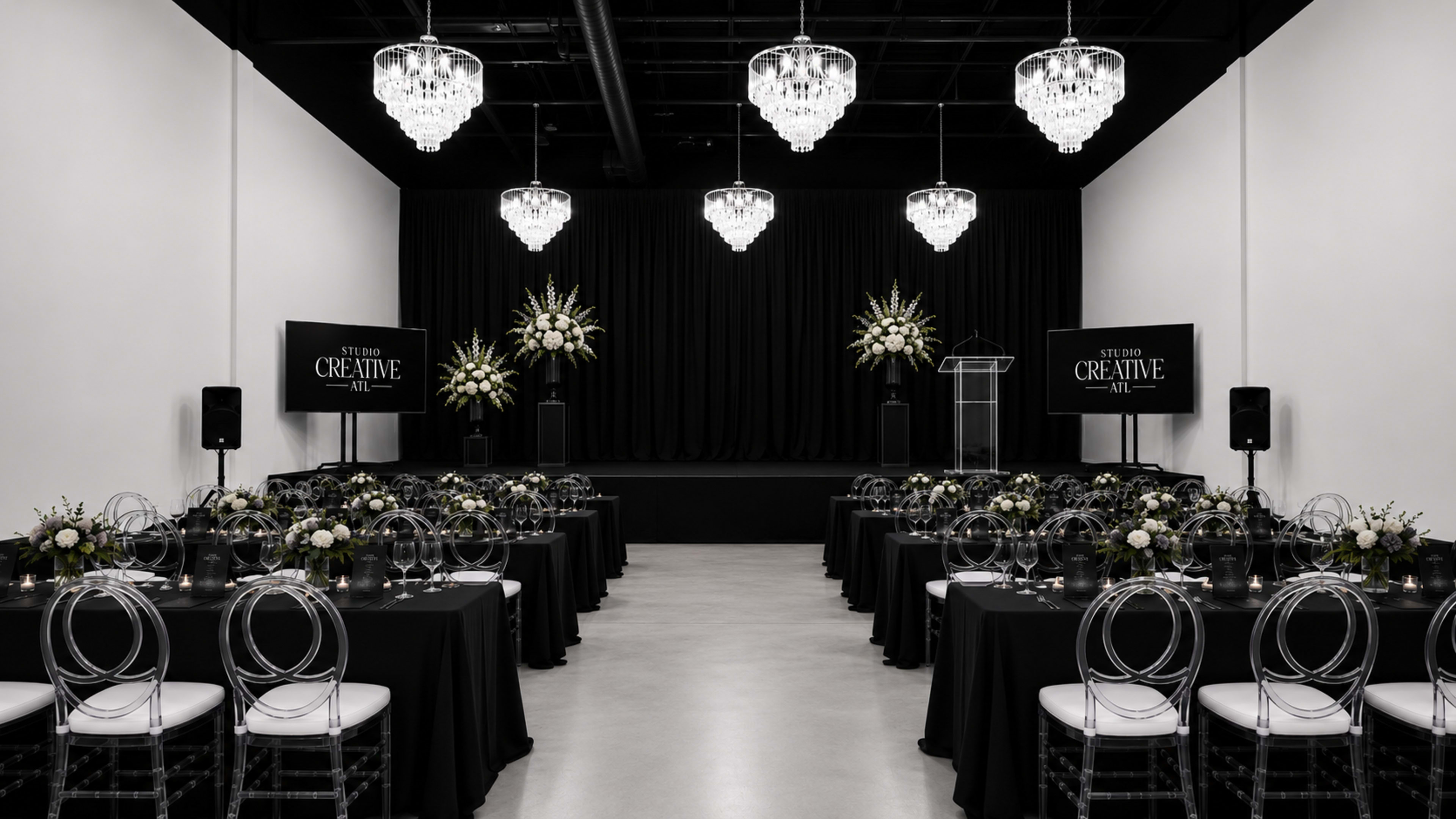 The image shows a banquet hall set up for an event, featuring rows of tables covered with black tablecloths, clear ghost chairs, and floral centerpieces, with a stage and screens at the front.