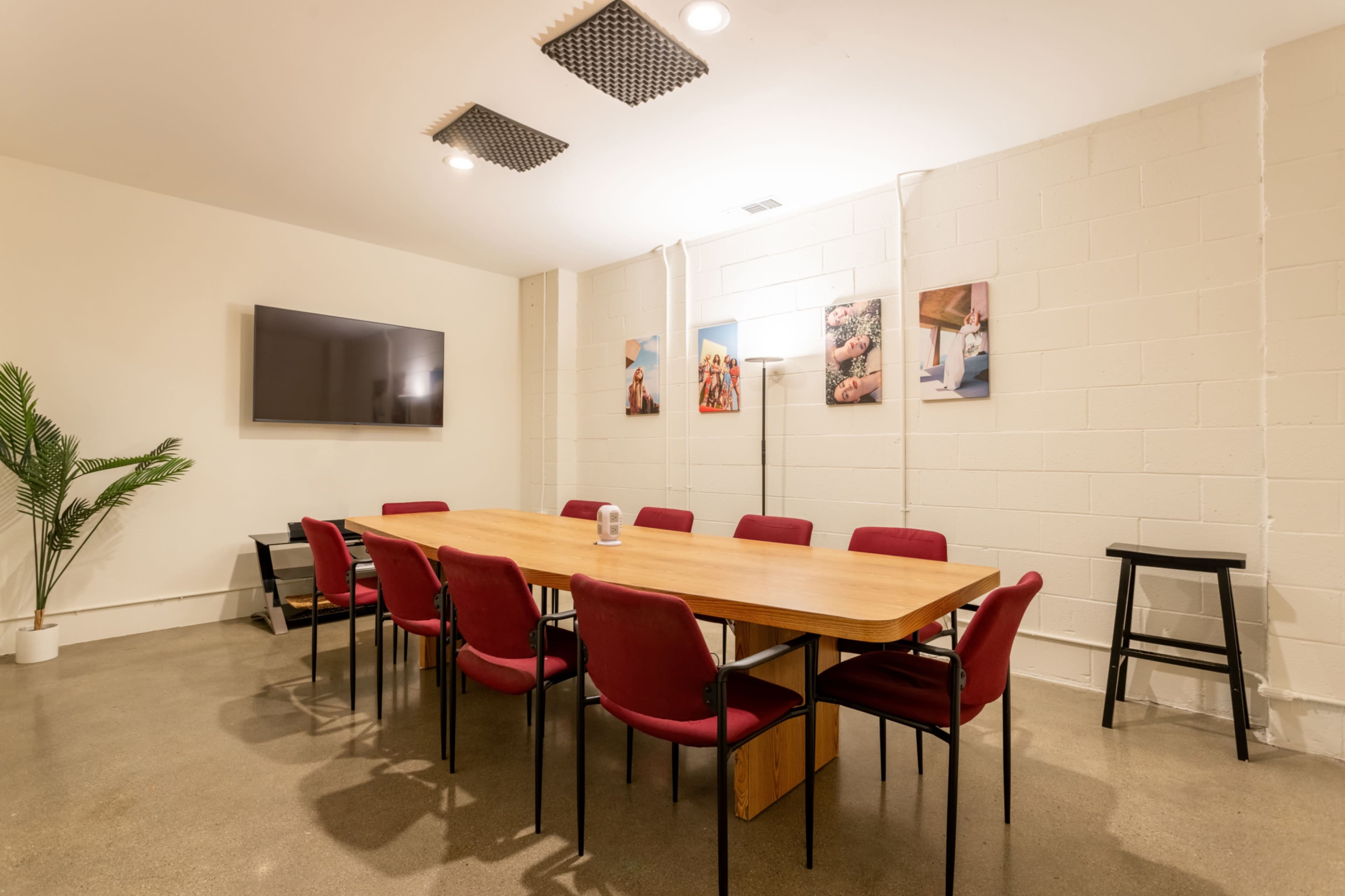 A long wooden table surrounded by red chairs is set up in a minimalist meeting room with a television on one wall and framed photos on another.