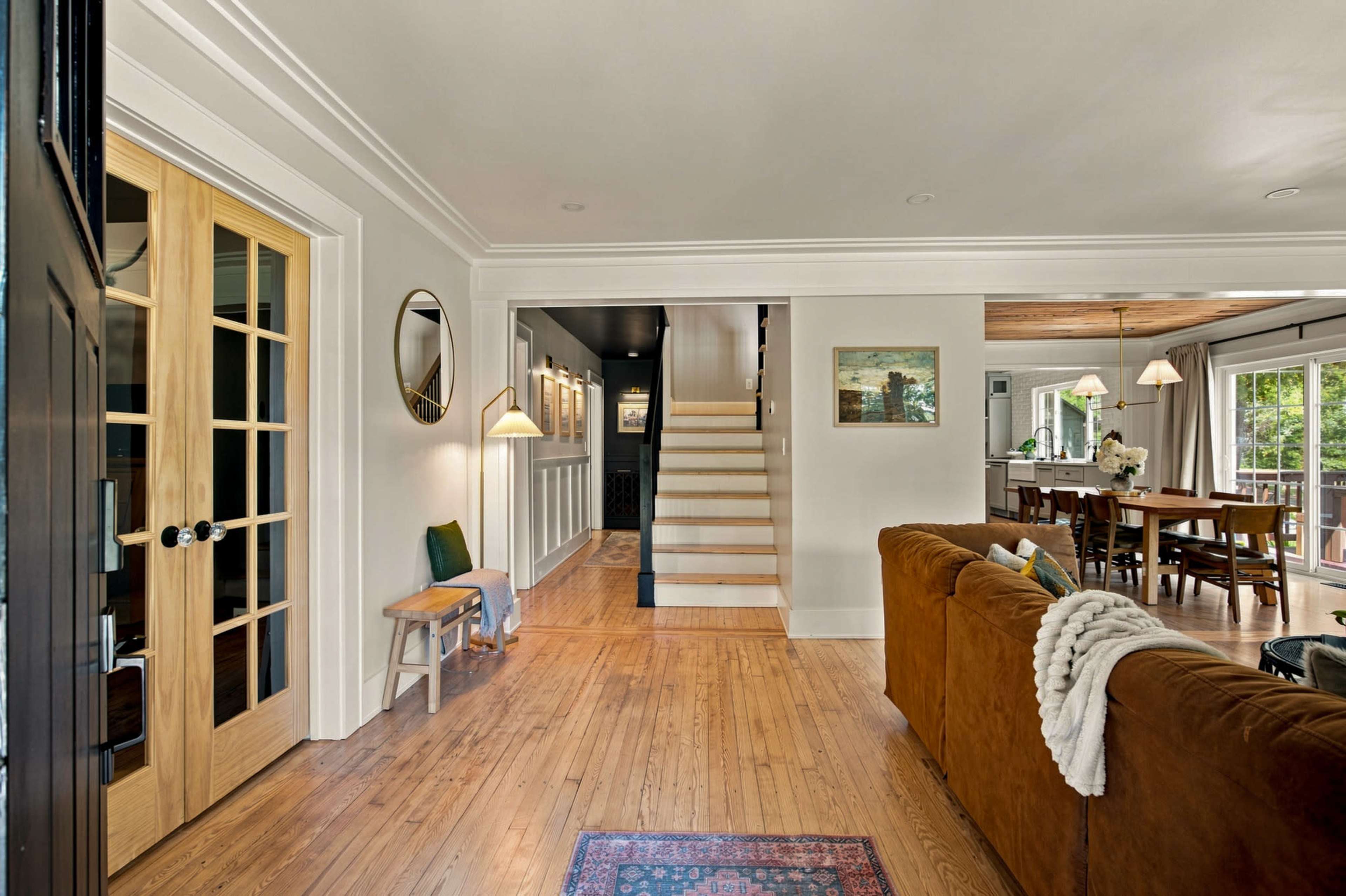 The image shows a well-lit entryway leading to a staircase and an open living space with a brown couch and wooden accents.