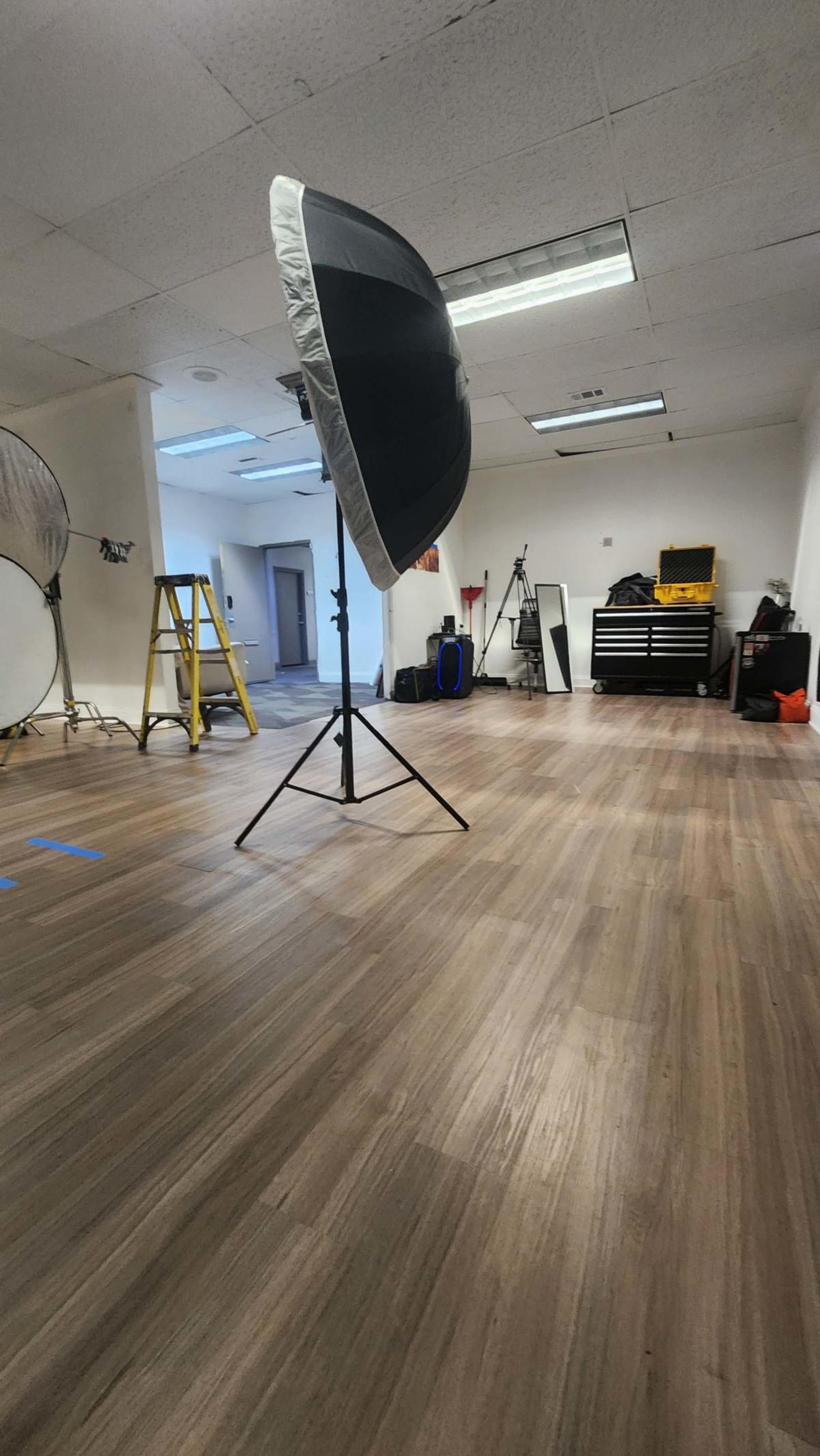 The image shows a spacious studio or room with wooden flooring, a large lighting umbrella on a stand, a ladder, and various equipment in the background.